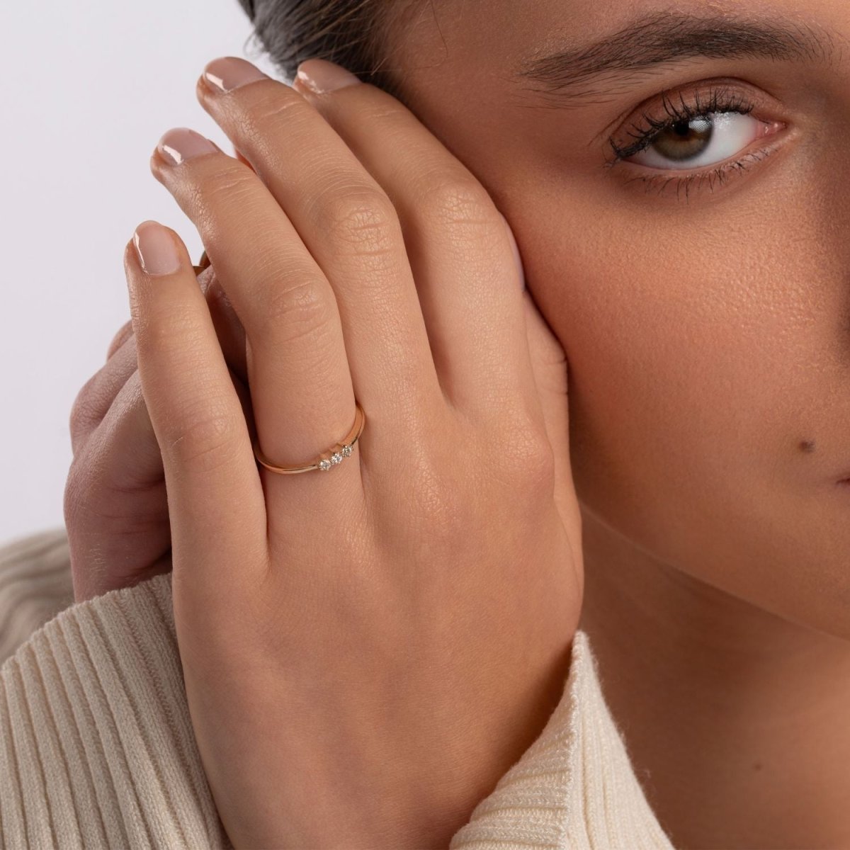 A close-up of a woman gently touching her face while wearing a 14k yellow gold three-stone diamond ring. The delicate design complements her refined and minimal style.
