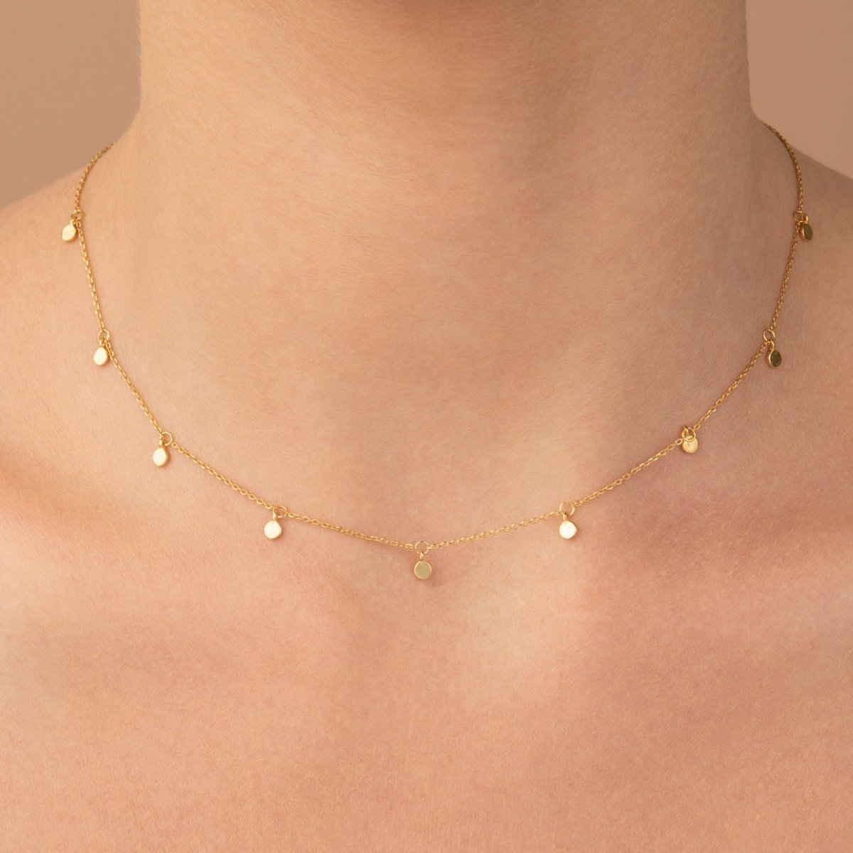 Woman wearing dainty yellow gold charm necklace – disc stations softly catching light on the collarbone.
