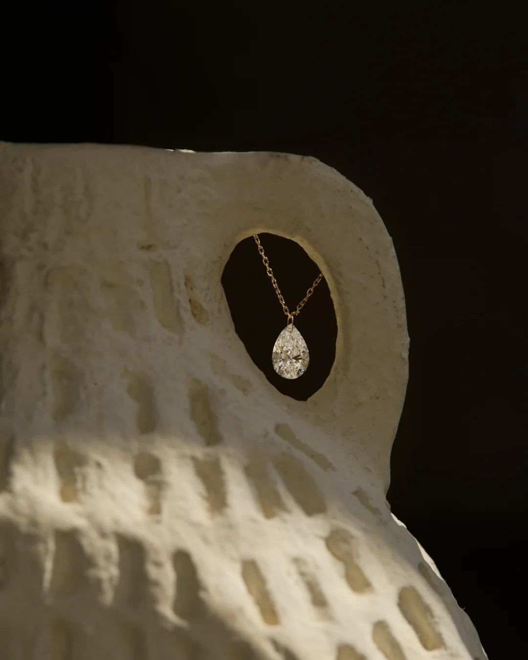Close-up of a pear-shaped diamond necklace suspended inside a ceramic sculpture, spotlighting fine gold craftsmanship under natural light.