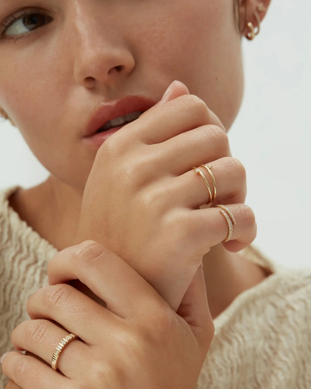 Close-up of a woman’s face and hands adorned with multiple diamond and gold rings, styled with soft knitwear and subtle gold earrings.