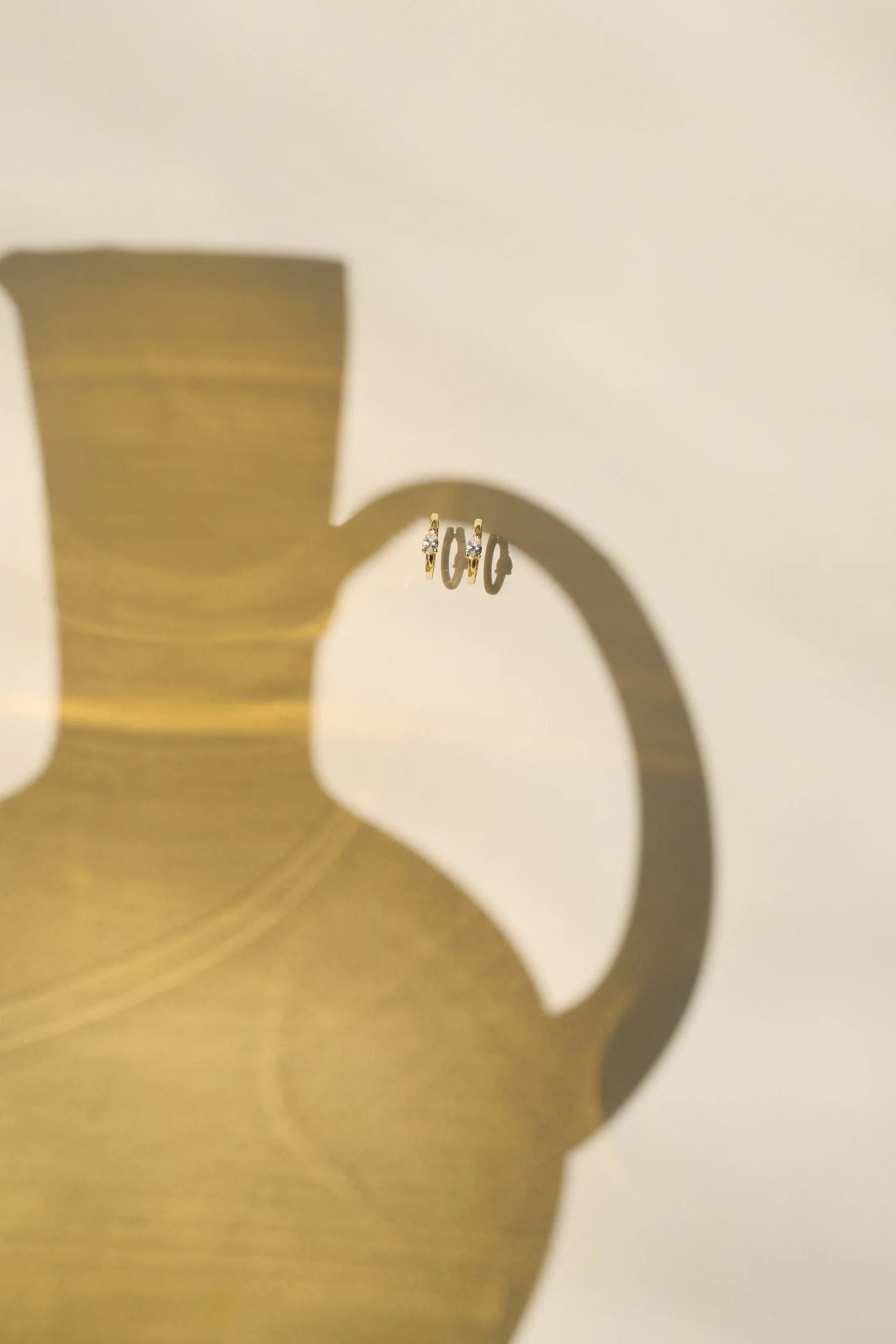 A delicate pair of gold huggie earrings adorned with a single diamond, suspended on the curved handle of a yellow ceramic jug, creating a dramatic shadow on a warm beige background.