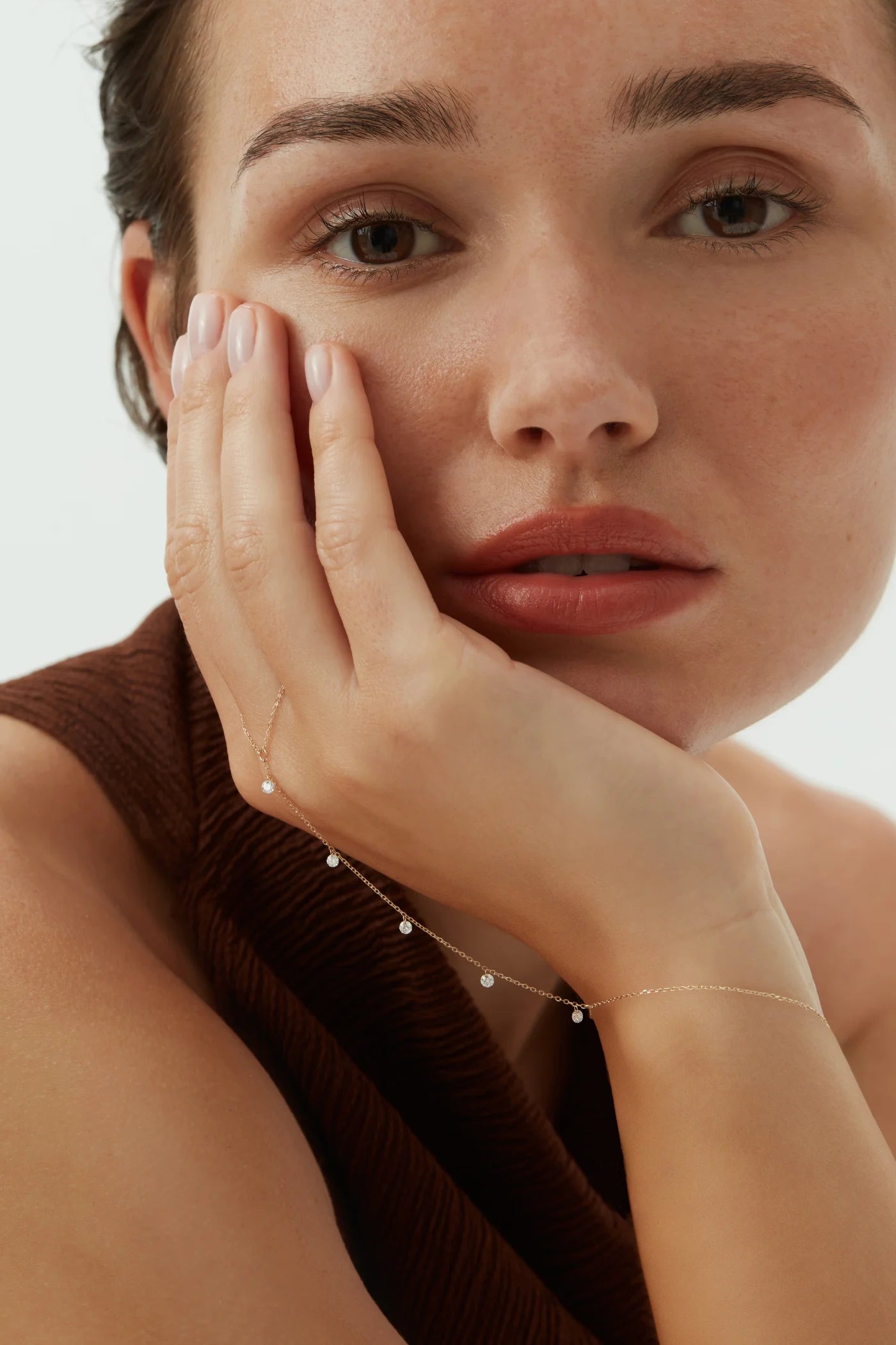 Close-up of a woman in a brown dress resting her hand on her face, wearing a delicate 14k yellow gold hand chain with five dangling diamonds.