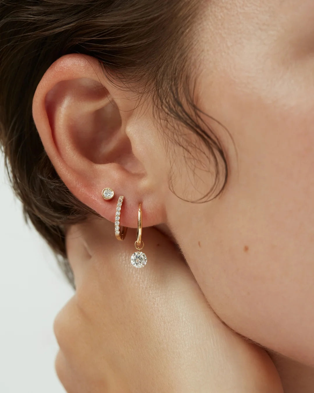 Close-up of a woman's ear adorned with three 14k solid gold earrings: a bezel-set diamond stud, a pavé diamond huggie, and a dangling solitaire diamond hoop.