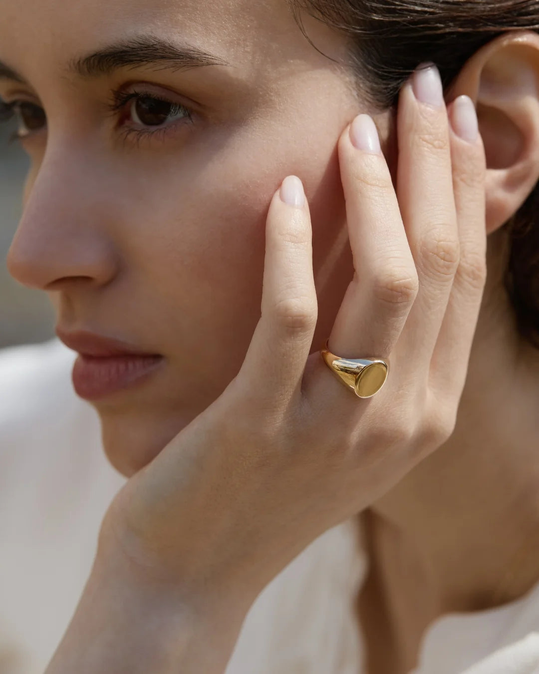 Woman with natural makeup resting her hand on her face, wearing a bold solid gold signet ring with a polished oval face.