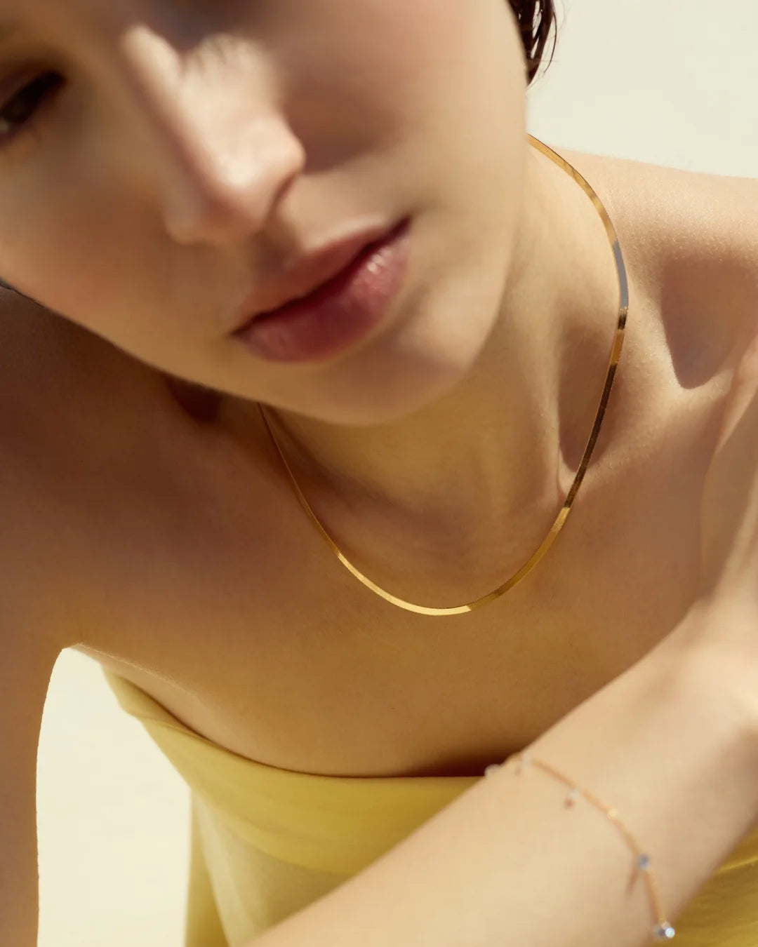 Close-up of a woman wearing a sleek gold flat chain necklace and a dainty diamond bracelet, styled against sunlit skin.