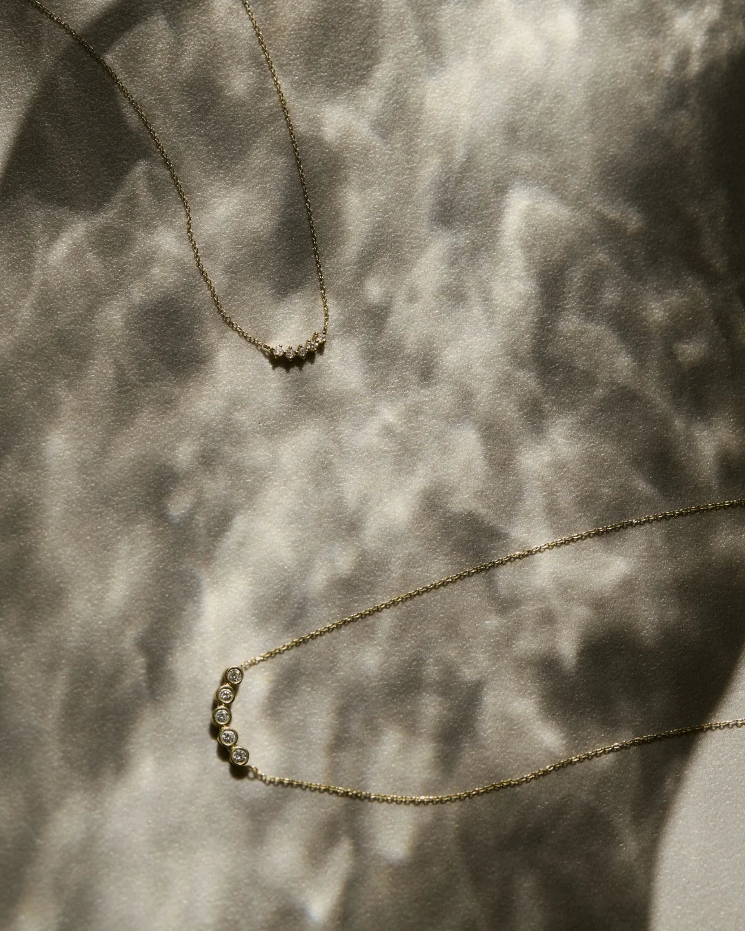 Two delicate diamond pendant necklaces displayed on a textured surface with soft natural light reflections.