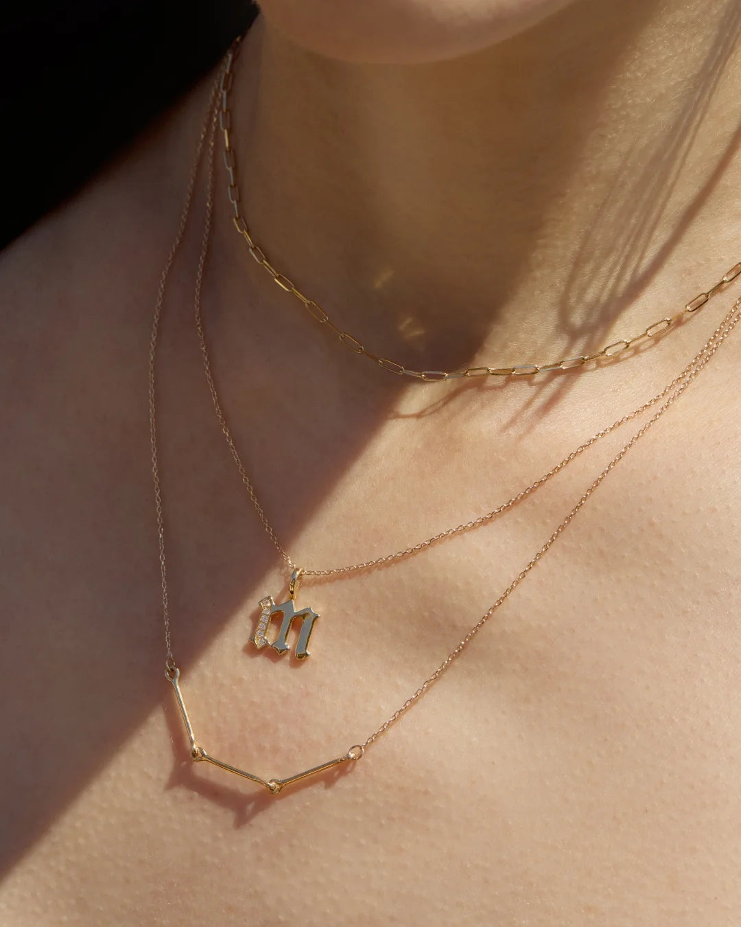 Minimal gold chains layered with zodiac and initial pendants resting on the collarbone, styled in soft sunlight.