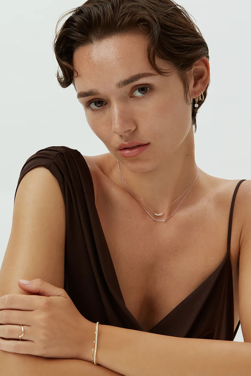 Model wearing the Iris Necklace in 14k rose gold, layered with another necklace and a matching gold bracelet, styled with a deep neckline top.