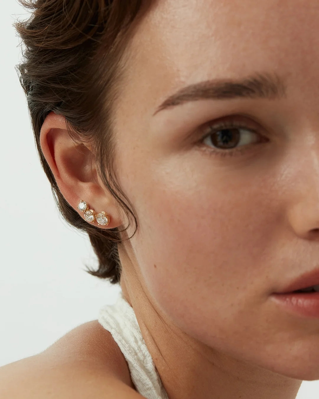 Close-up of a woman’s ear adorned with three round-cut diamond stud earrings set in yellow gold, styled with a white textured halter top.