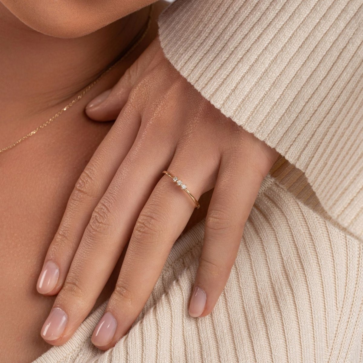 A woman wearing a 14k yellow gold three-stone diamond ring, resting her hand on a soft sweater. The round diamonds catch the light, adding subtle elegance.