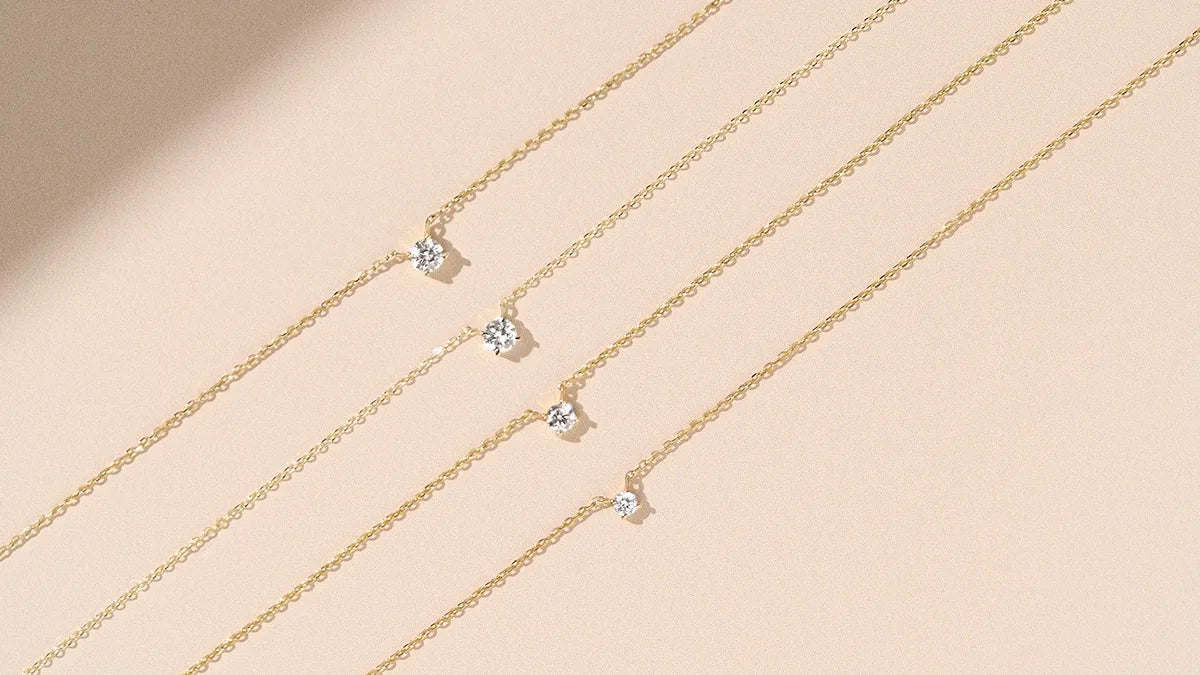 Three 14k gold diamond station necklaces with prong-set lab grown diamonds arranged diagonally on a warm beige surface with natural shadow | Enea Studio