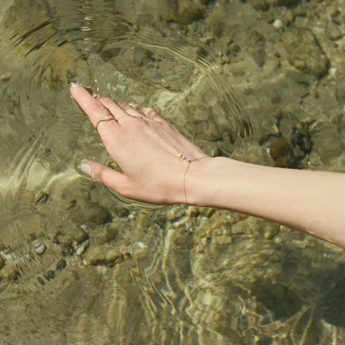 A woman&#39;s hand styled with a dainty 14k hand chain diamond bracelet and a wavy diamond band subtly touching still water creating soft ripples-Enea