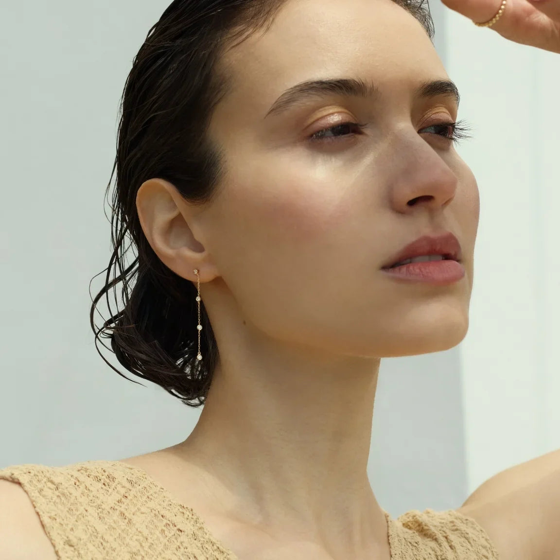 Side profile of a woman with wet, slicked-back hair wearing a 14k gold drop earring featuring four evenly spaced diamonds along a delicate chain, set against a soft beige background-Enea
