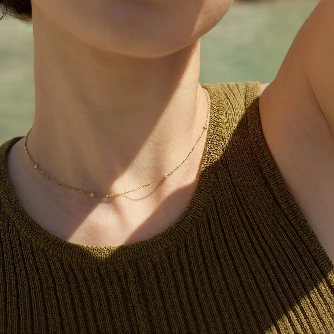 Close up of a woman in olive green dress wearing a dainty 14k gold necklace adorned with 5 tiny diamonds scattered evenly across the chain, glinting softly under soft natural light-Enea
