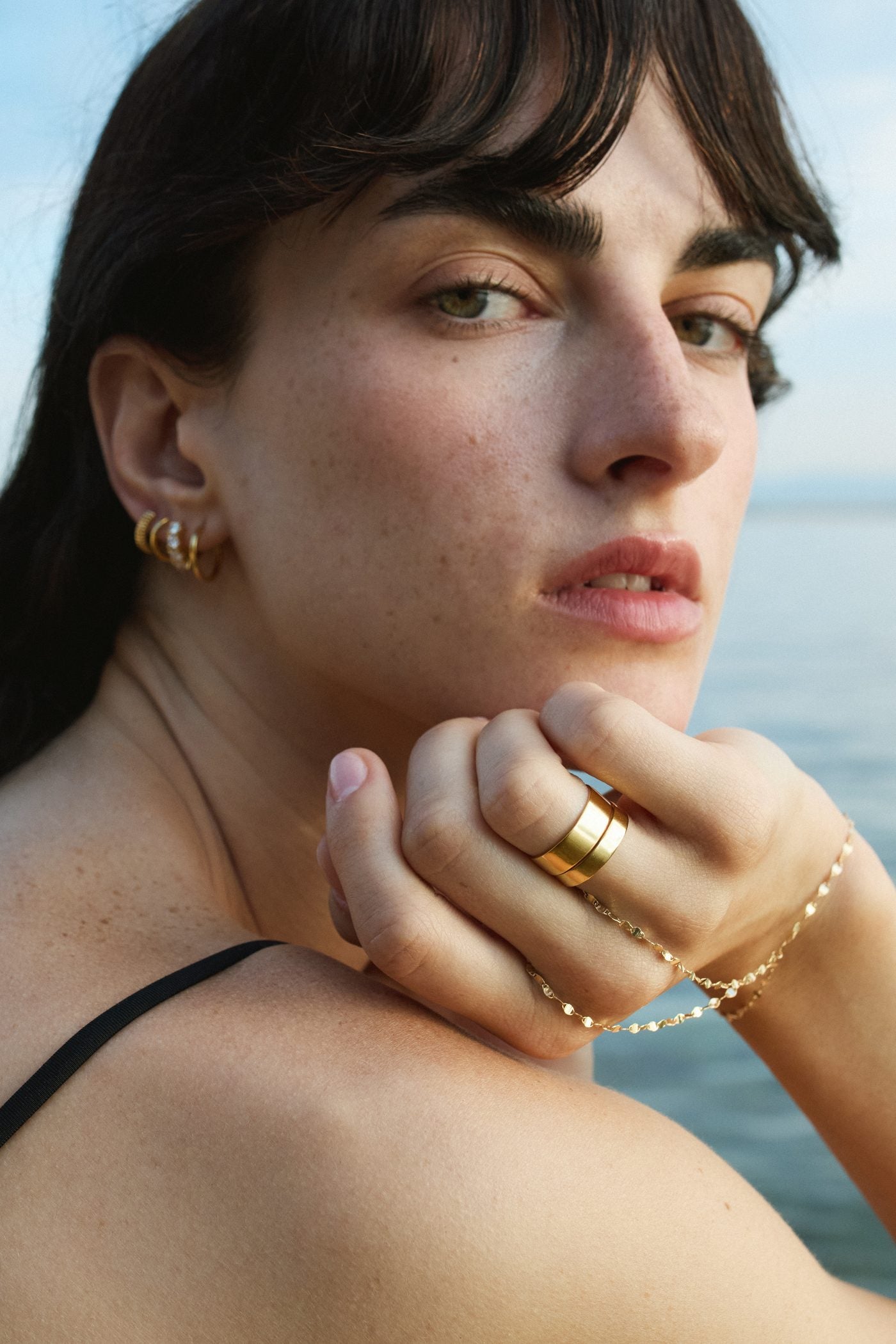 Model wearing solid gold hoop earrings, stacked rings and chain bracelets by the sea