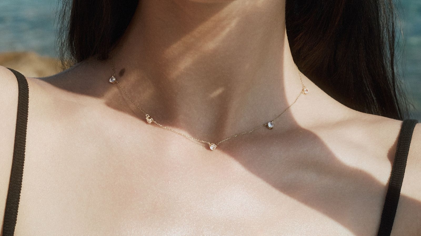 Close-up of a woman's neck under soft natural light styled with a solid gold diamond station necklace by ENEA against a blurred sea background