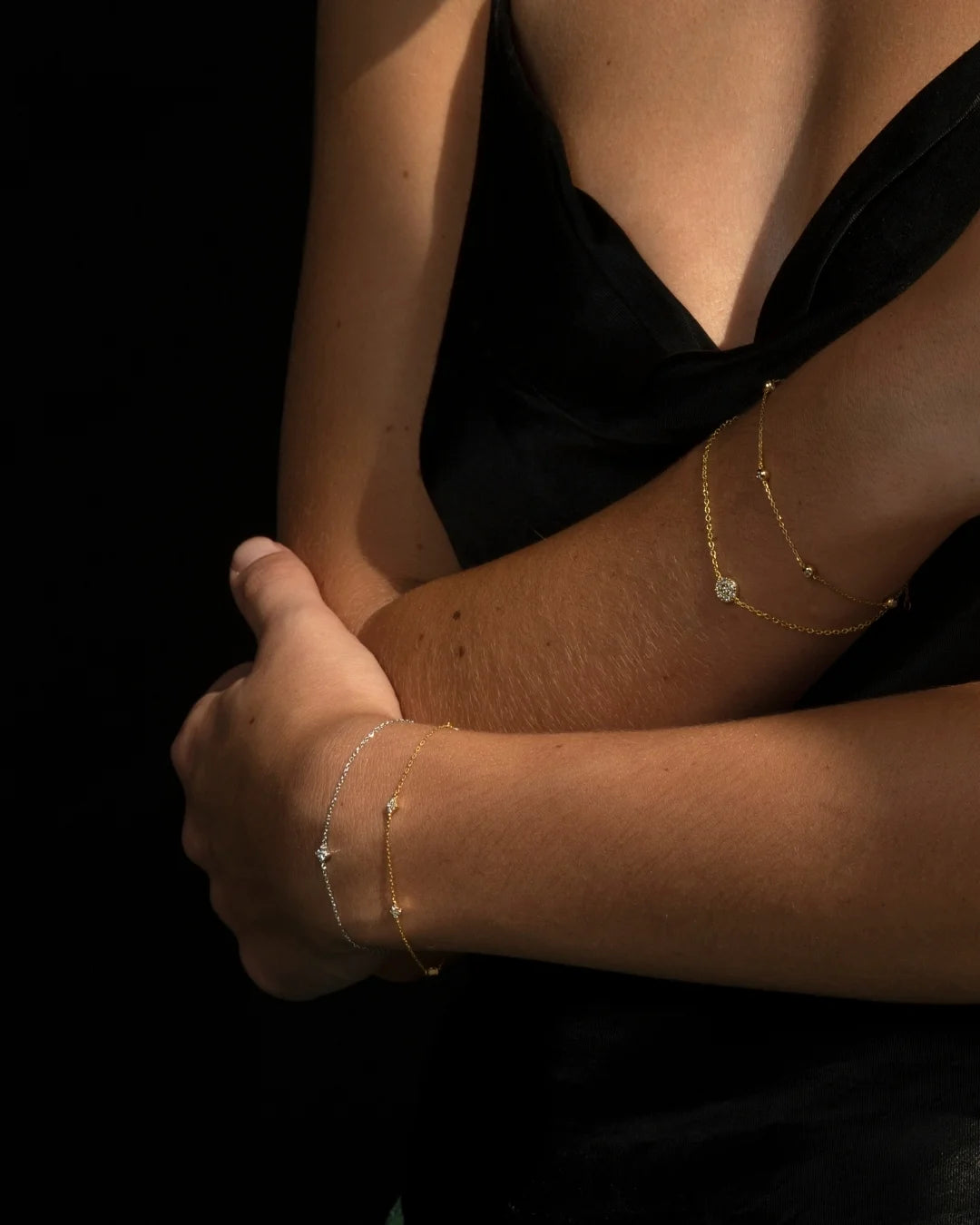 Close-up of a woman's arms styled with dainty 14k gold diamond bracelets shining under soft sunlight, against a black background. Enea Studio