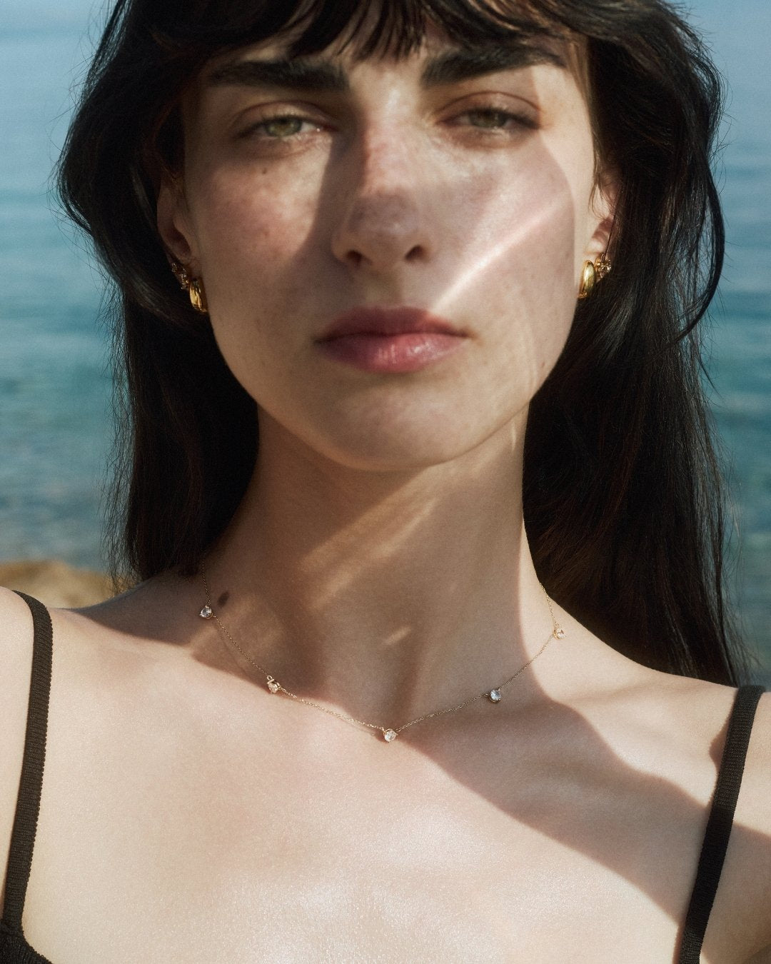 Woman wearing a solid gold station necklace with 5 diamonds and multiple gold earrings by ENEA under soft natural light on a blurred sea background