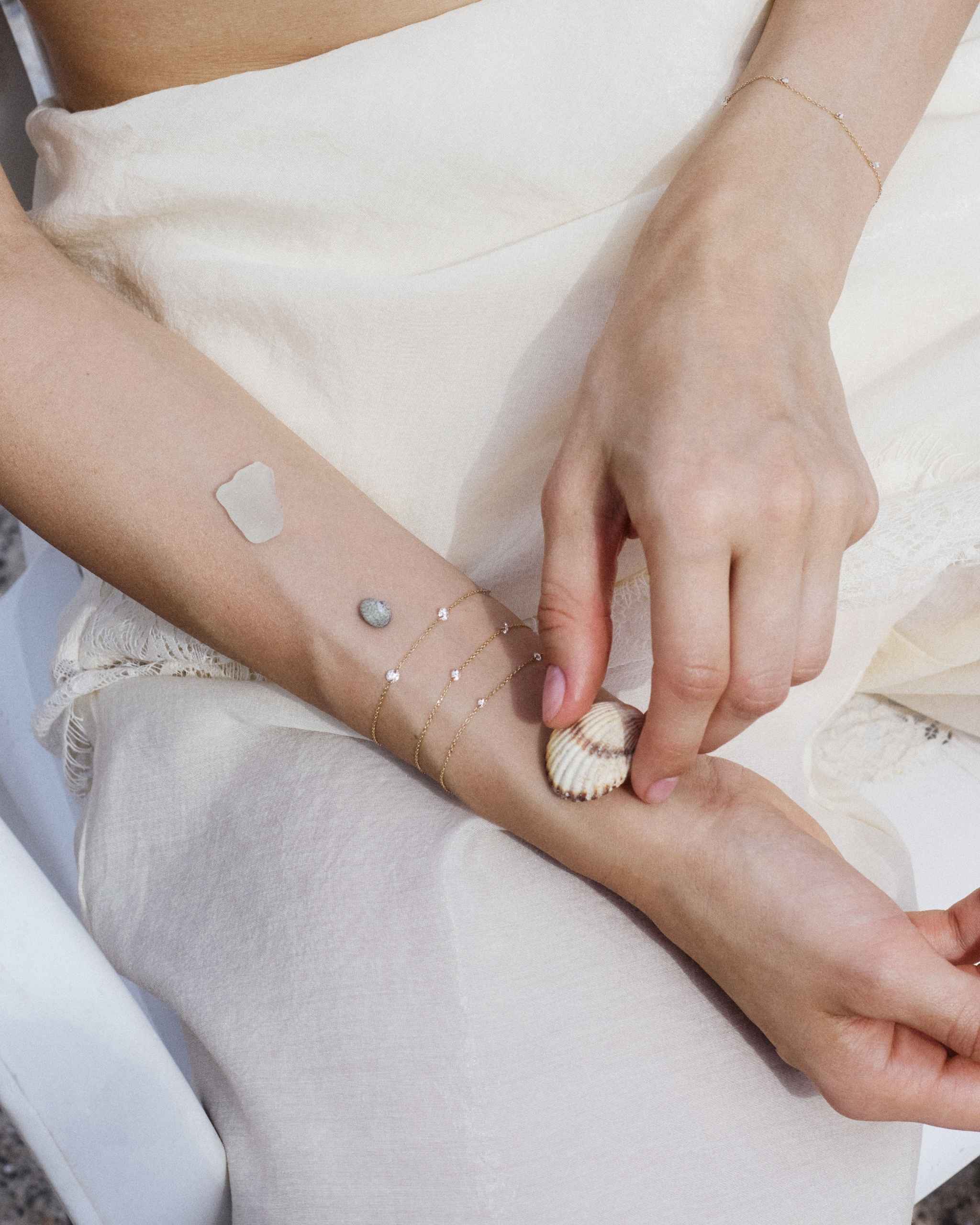 a woman laying her wrist on her leg styled with three gold chain bracelets with delicate nude diamonds of different sizes next to small sea shells