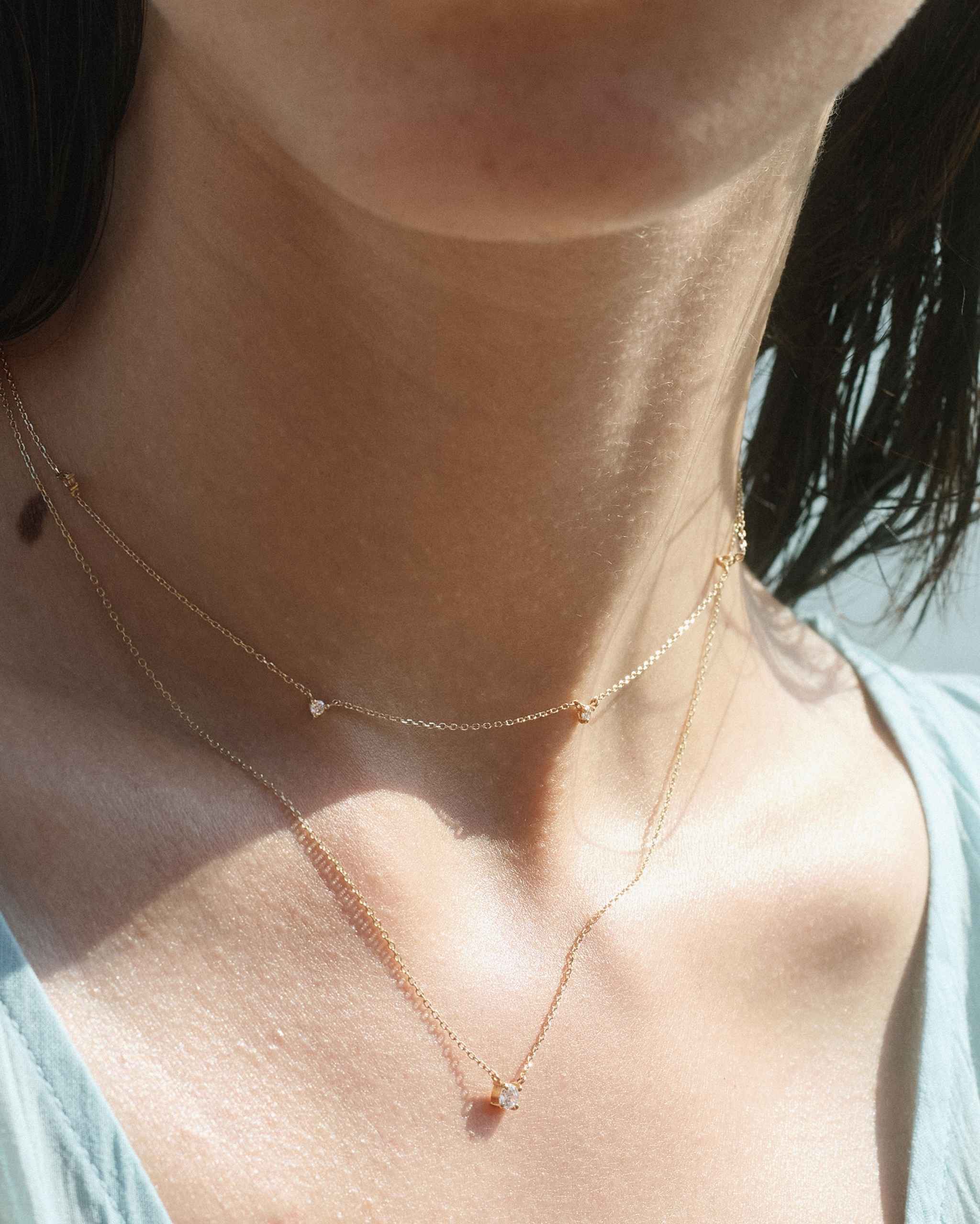 Close up of a woman's neck wearing a solid gold diamond by the yard necklace with five diamonds layered with a petite solid gold diamond solitaire necklace under soft natural light
