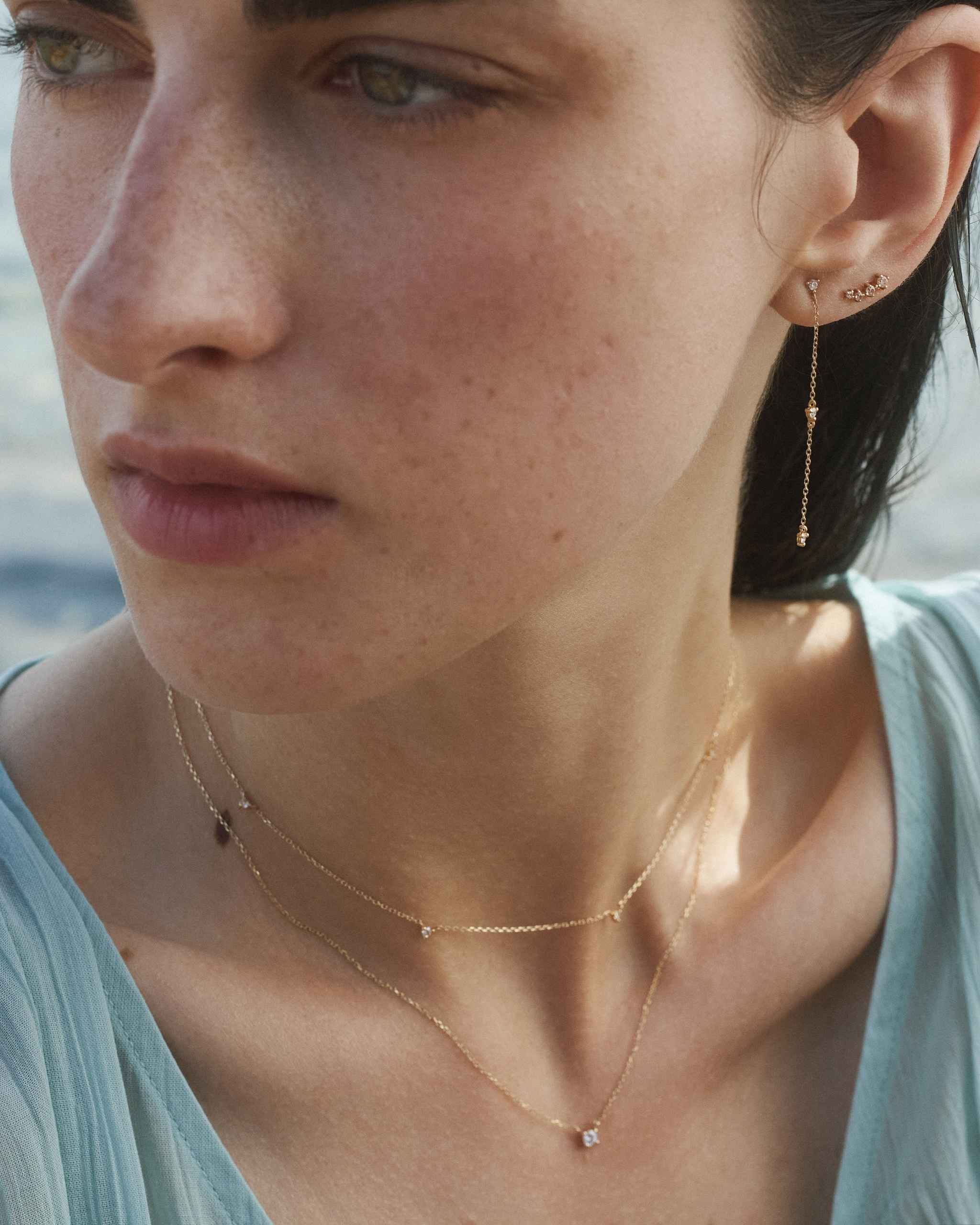 woman wearing a solid gold diamond by the yard necklace with five diamonds layered with a petite solid gold diamond solitaire necklace, and a gold chain earring with three dainty diamonds stacked with a curved diamond stud