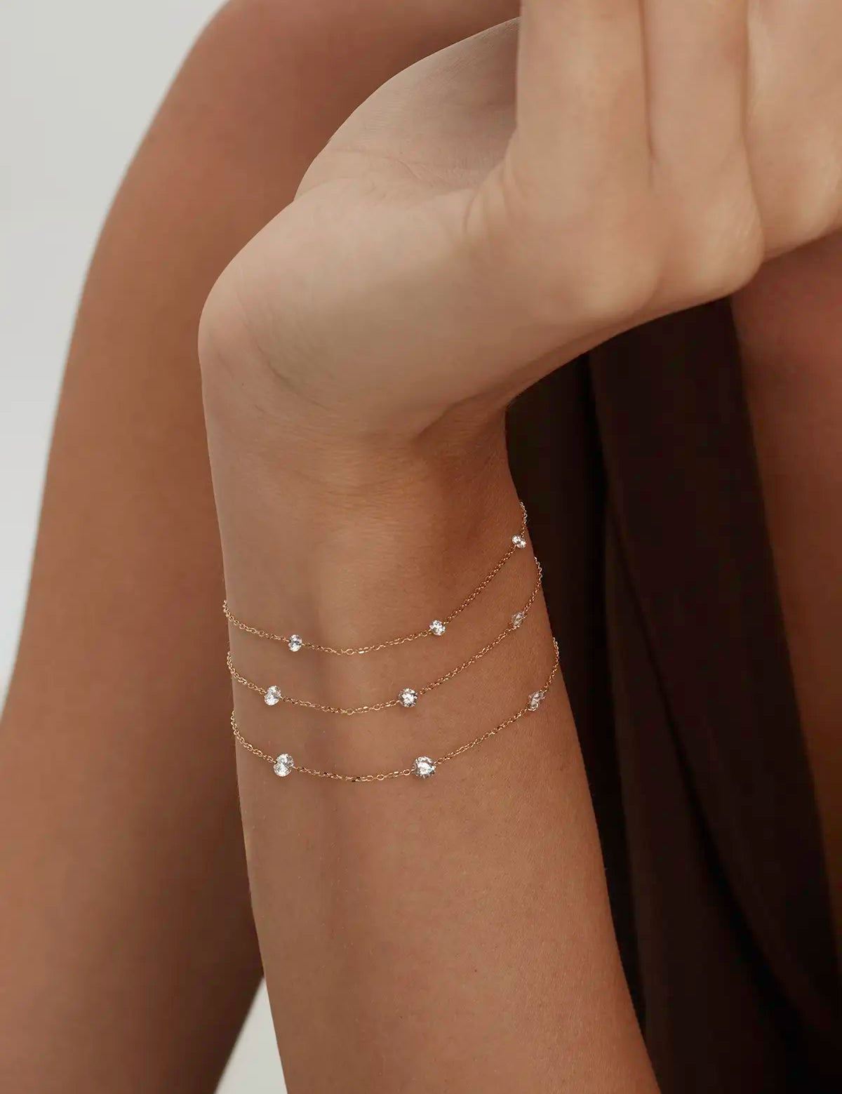 Close-up of a woman's wrist wearing three delicate gold bracelets with dainty, floating diamonds on a neutral background. Enea Studio