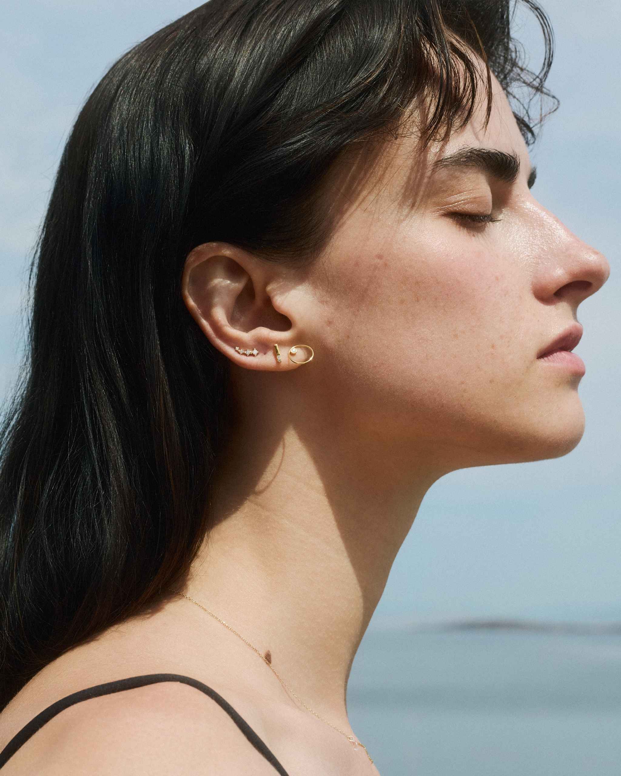 profile view of a woman wearing small gold studs with delicate diamonds on a blurred sea background