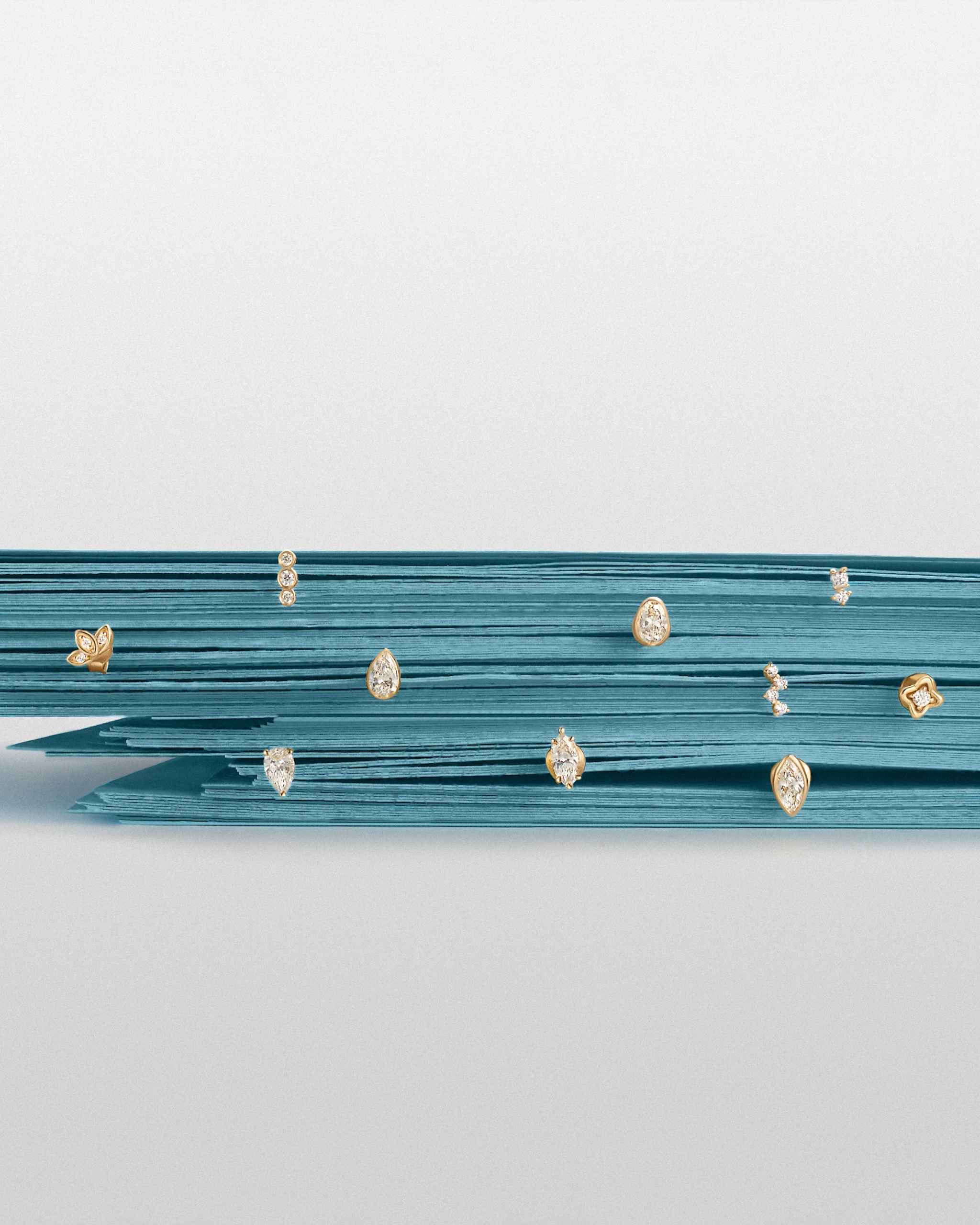 A stack of teal paper sheets displaying multiple small gold and diamond stud earrings with different sizes and shapes, set against a light gray background.