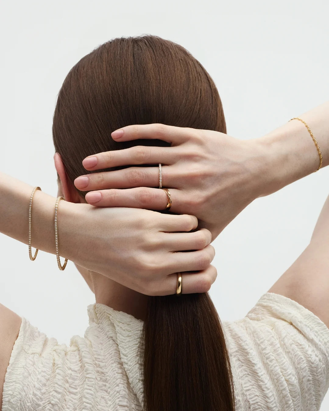 Back view of a woman with long brown hair holding a sleek ponytail with her hands which are styled with gold rings and dainty diamond tennis bracelets on her wrists. Enea Studio