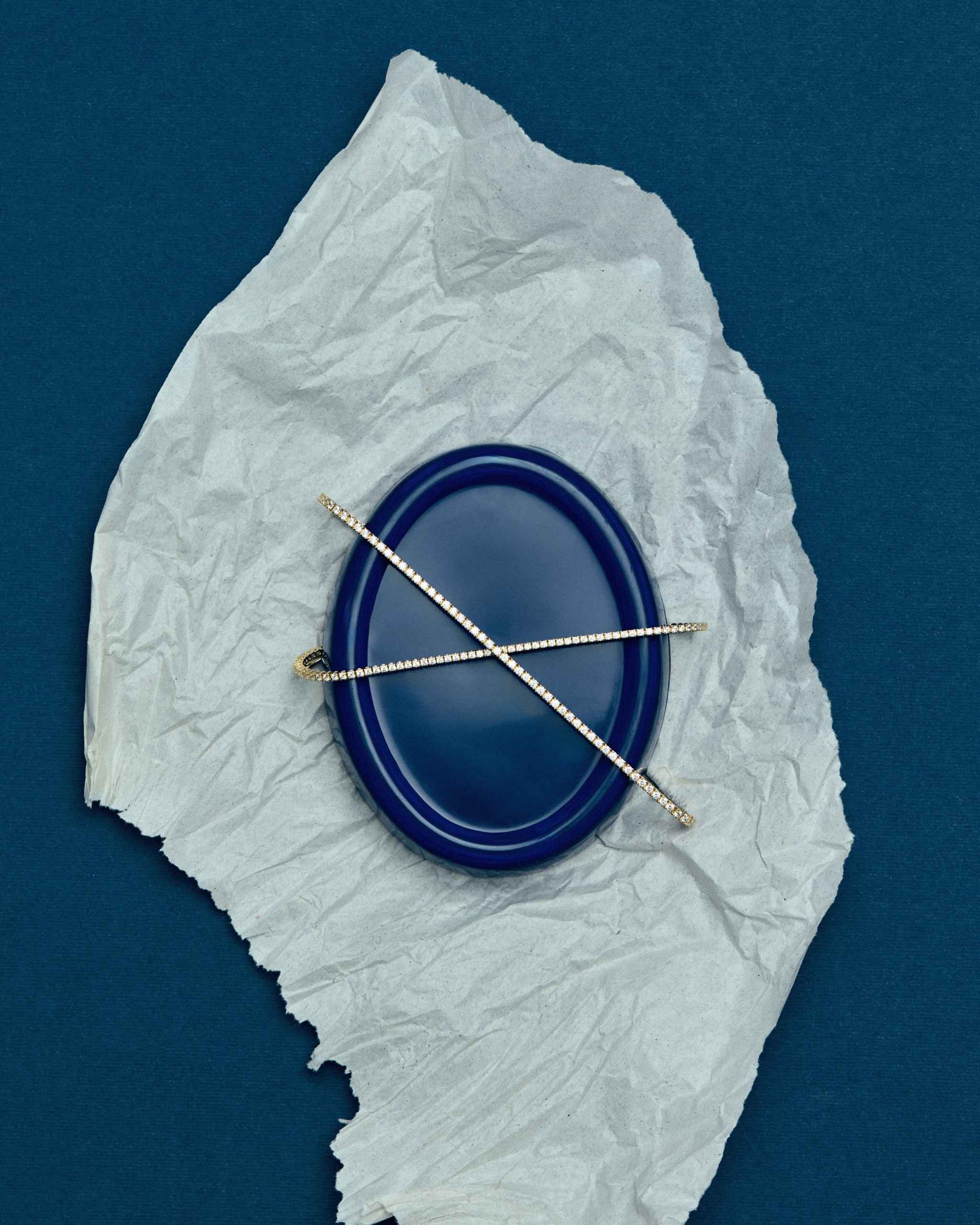 Two gold diamond tennis bracelets crossed in an X shape on a blue plate on crumpled white paper against a blue background