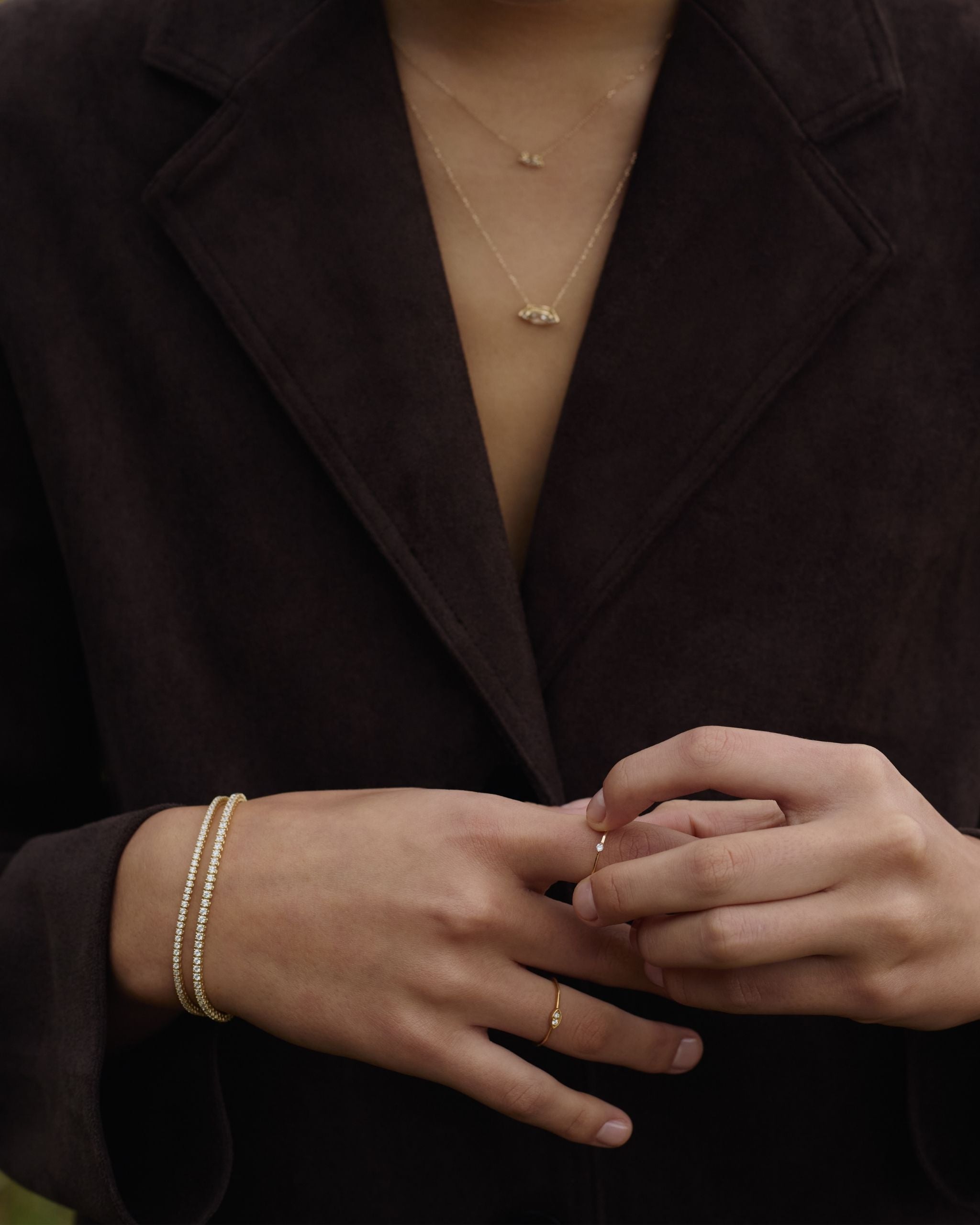 woman in brown jacket wears 14k yellow gold diamond tennis bracelets and thin gold rings with delicate diamonds