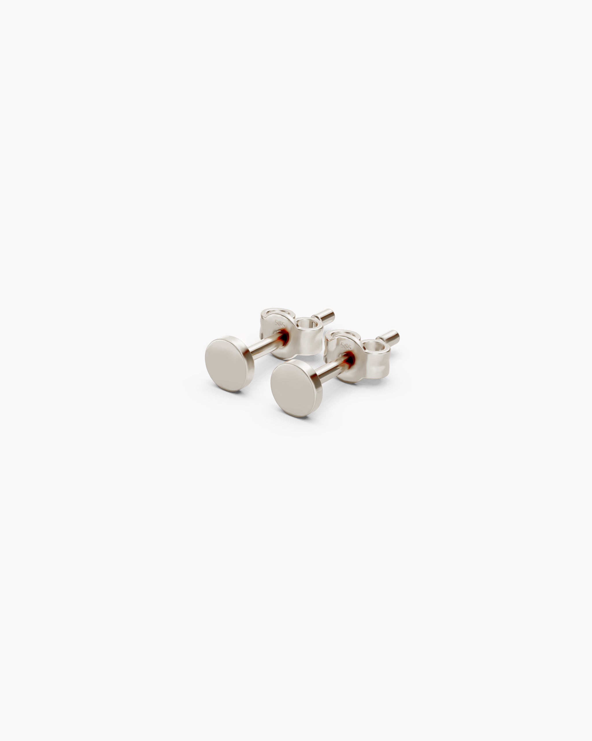 Angled view of the Diskos Disc Studs in 14k white gold with push back closures stamped 585, on an off-white surface. ENEA Studio