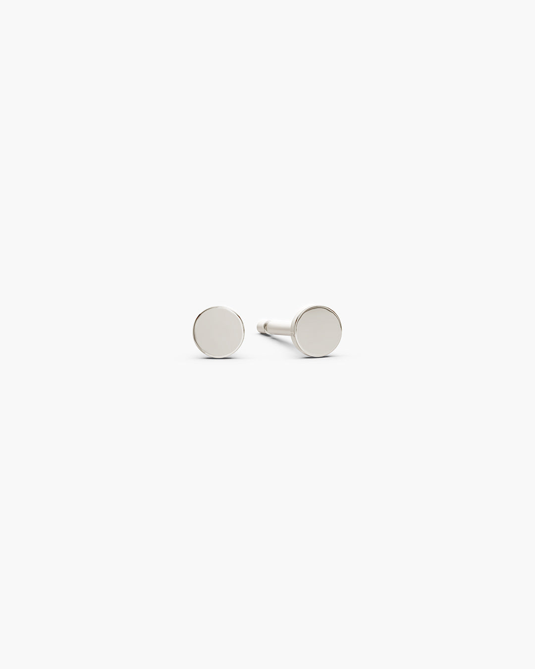 Diskos Disc Studs in 14k white gold, front view, a pair of 3.5mm polished flat white gold disc stud earrings on an off-white surface. ENEA Studio