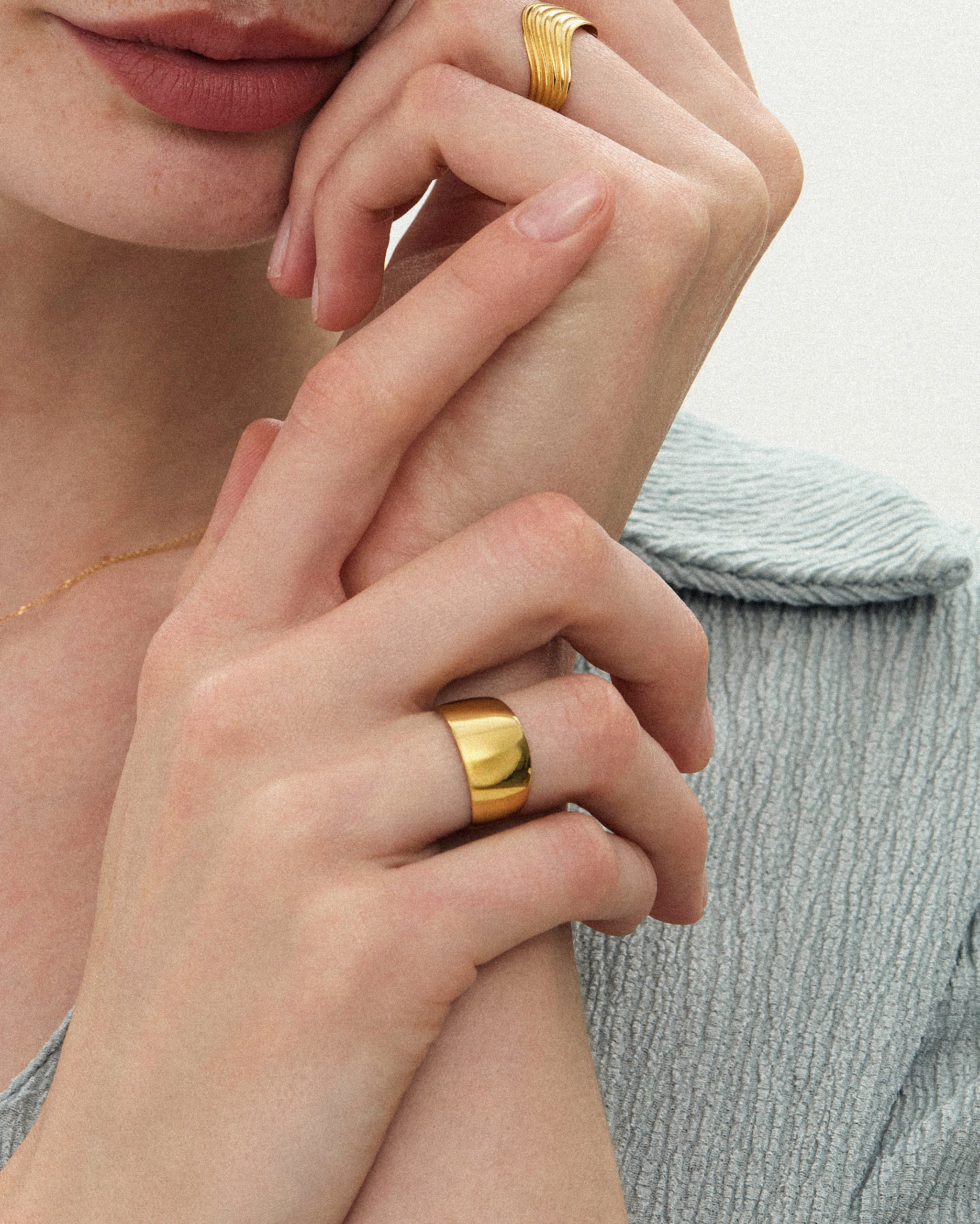 Close-up of a woman's hands styled with a wavy ribbed solid gold band and a wide, polished yellow gold band on a neutral background