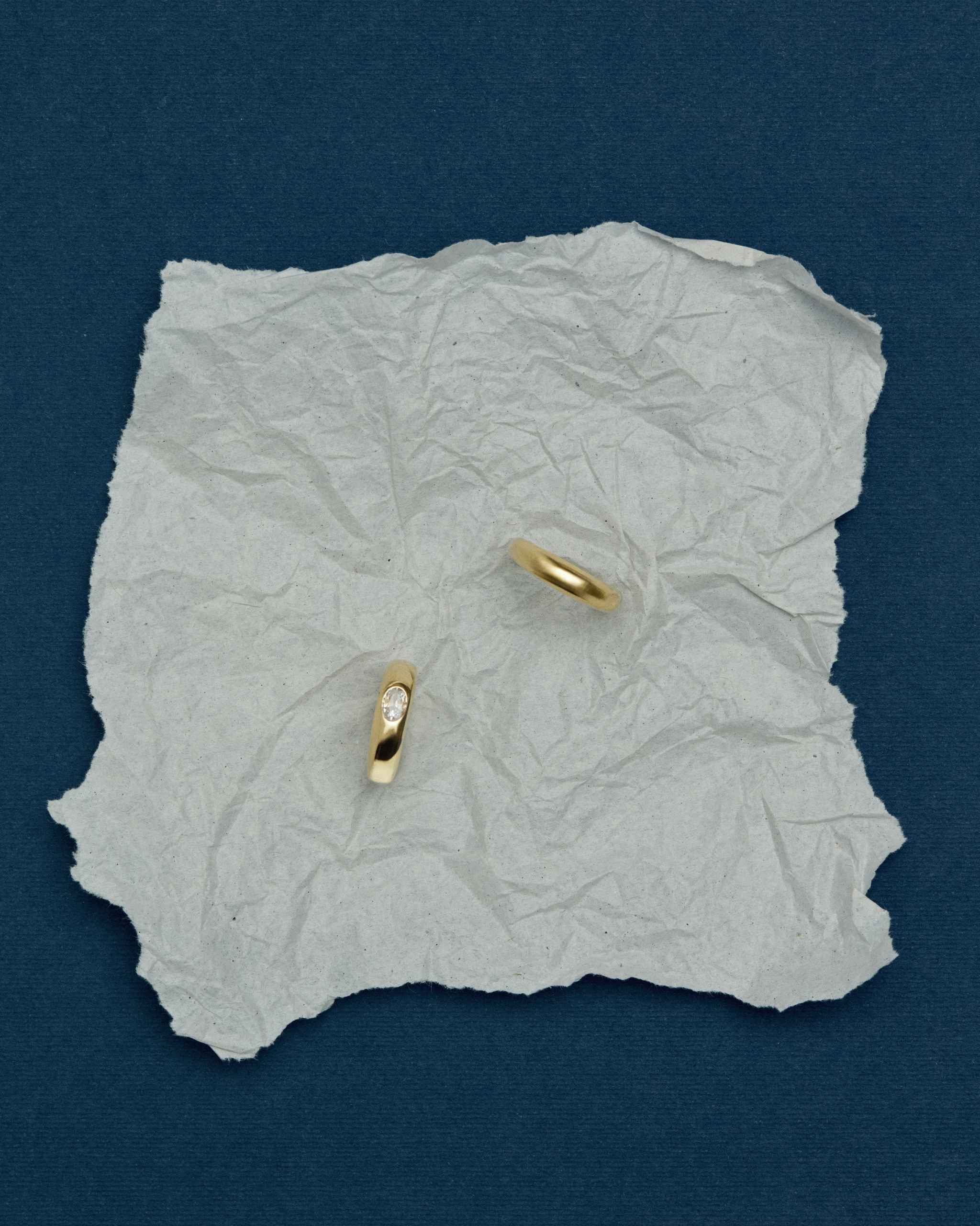 two solid gold dome rings, one plain and one set with a round diamond at its center, placed on a crumpled piece of paper on a blue surface