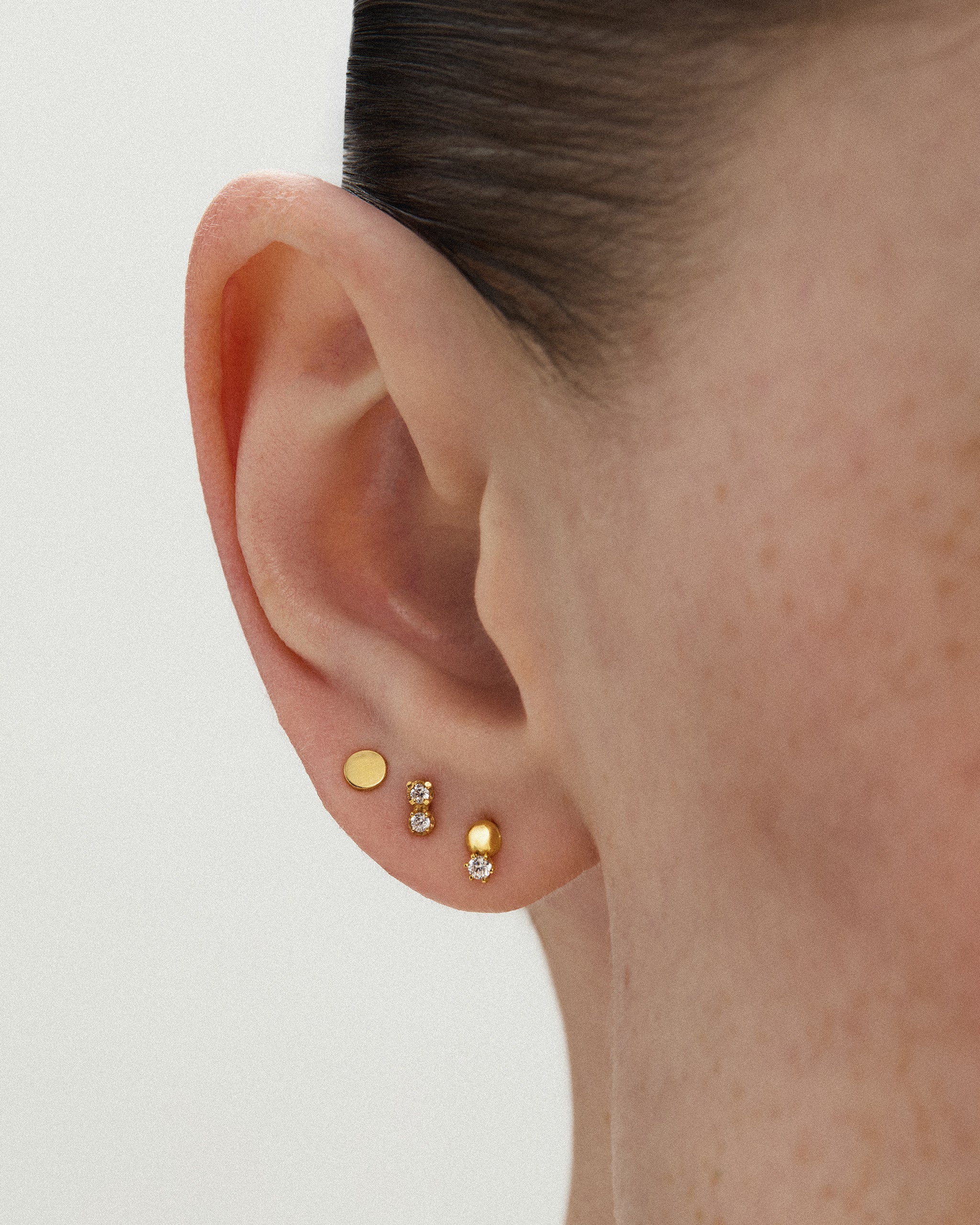 Close up of a woman's ear wearing the Dyo Diamond Bead Stud in 14k yellow gold in the lobe, worn bead up, stacked with two small gold studs. ENEA Studio