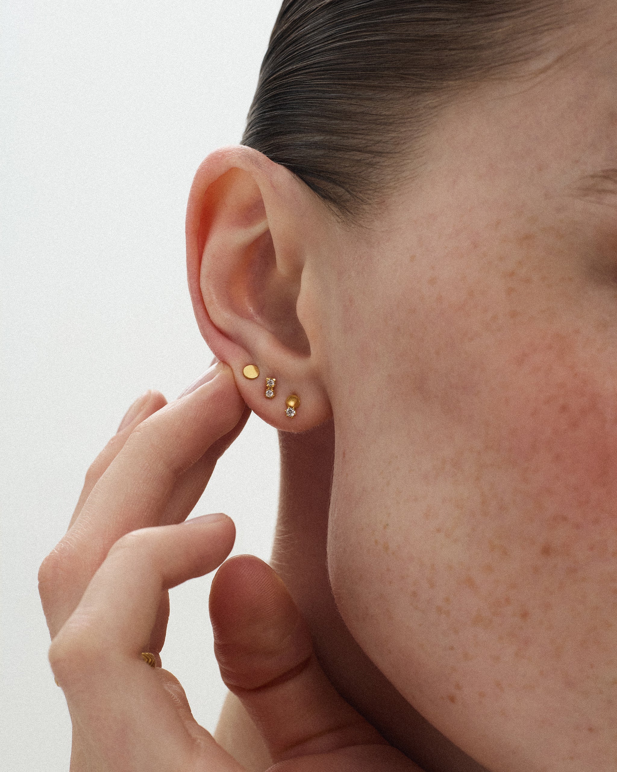 Woman touching her ear stacked with three 14k yellow gold studs including the Dyo Diamond Bead Stud, a lab grown diamond beside a gold bead. ENEA Studio