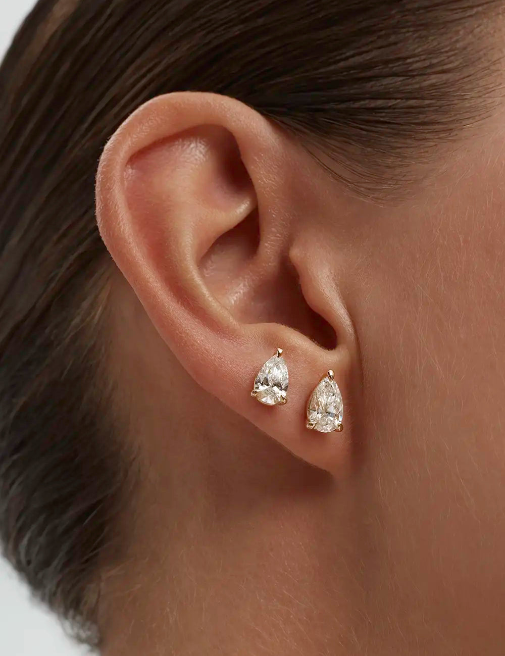Close-up of a woman's ear wearing two pear-shaped diamond solitaire earrings against a neutral background. Enea Studio
