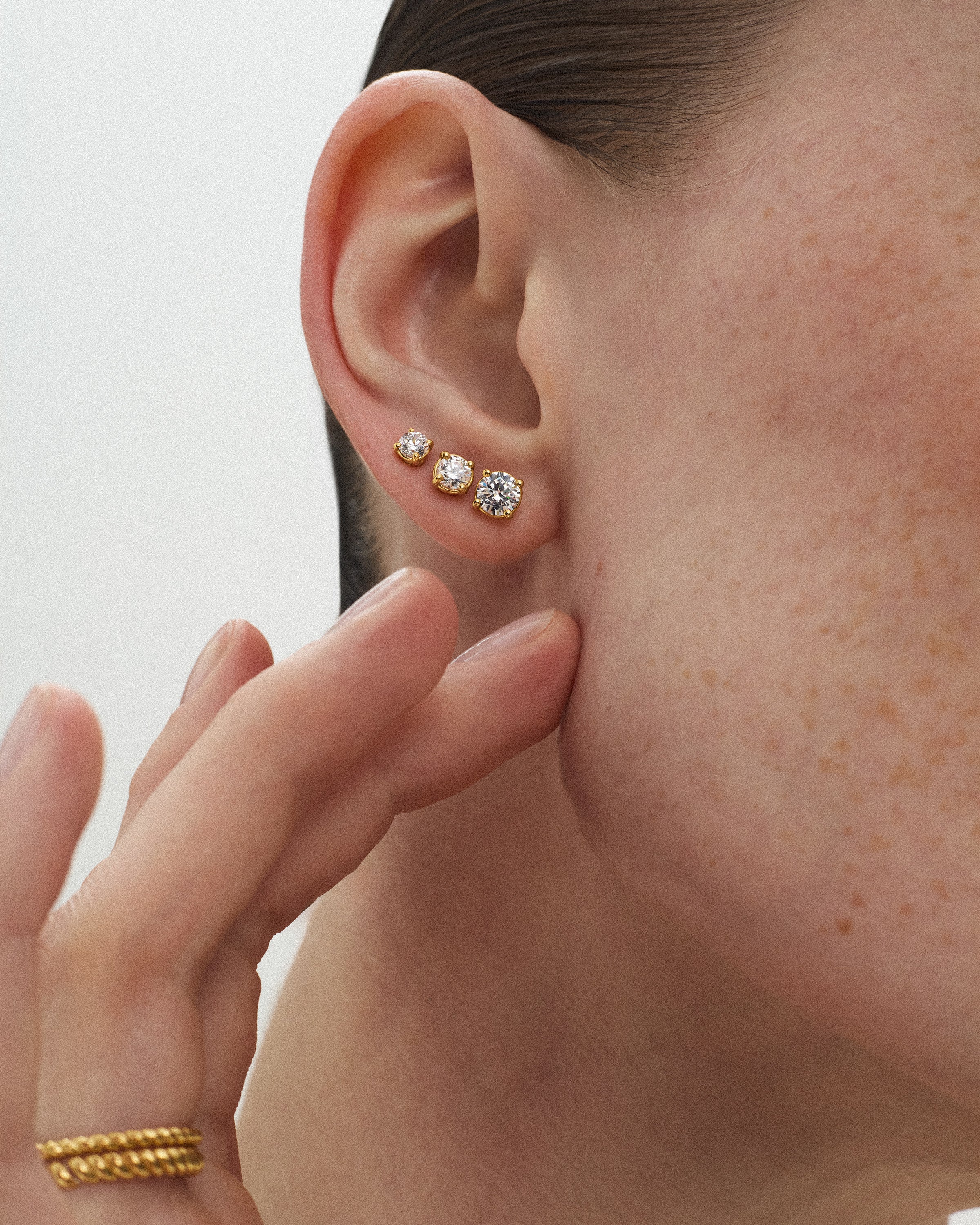 A close-up of a woman's ear wearing three eclat grand stud earrings of different diamond sizes in 14k yellow gold against a light off-white background.