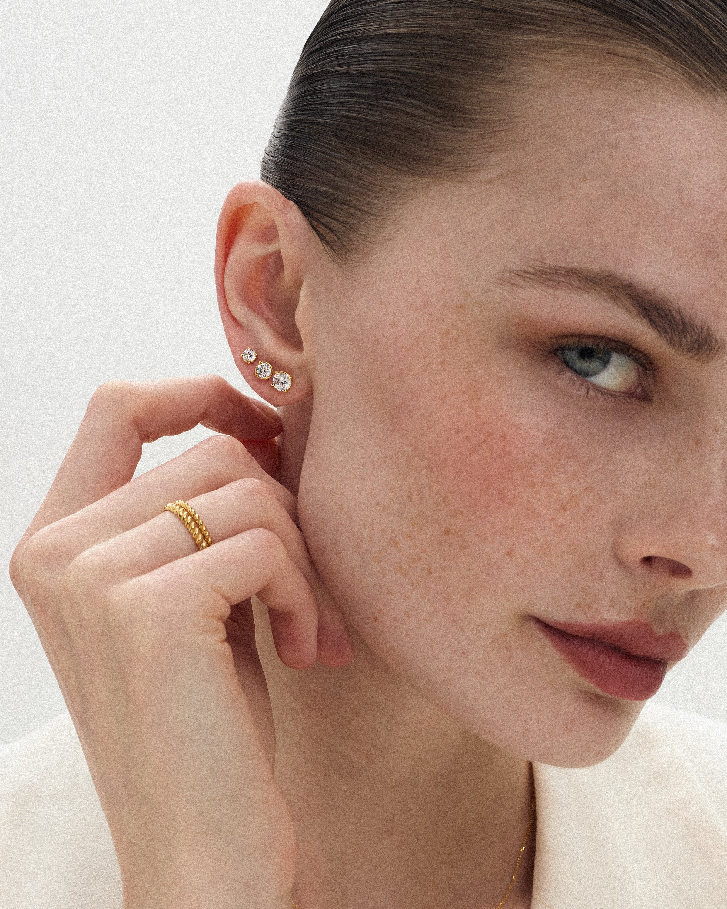Angled view of a woman wearing 14k yellow gold bands alongside three eclat grand diamond studs of slightly different sizes with her hand touching her neck against a soft light background