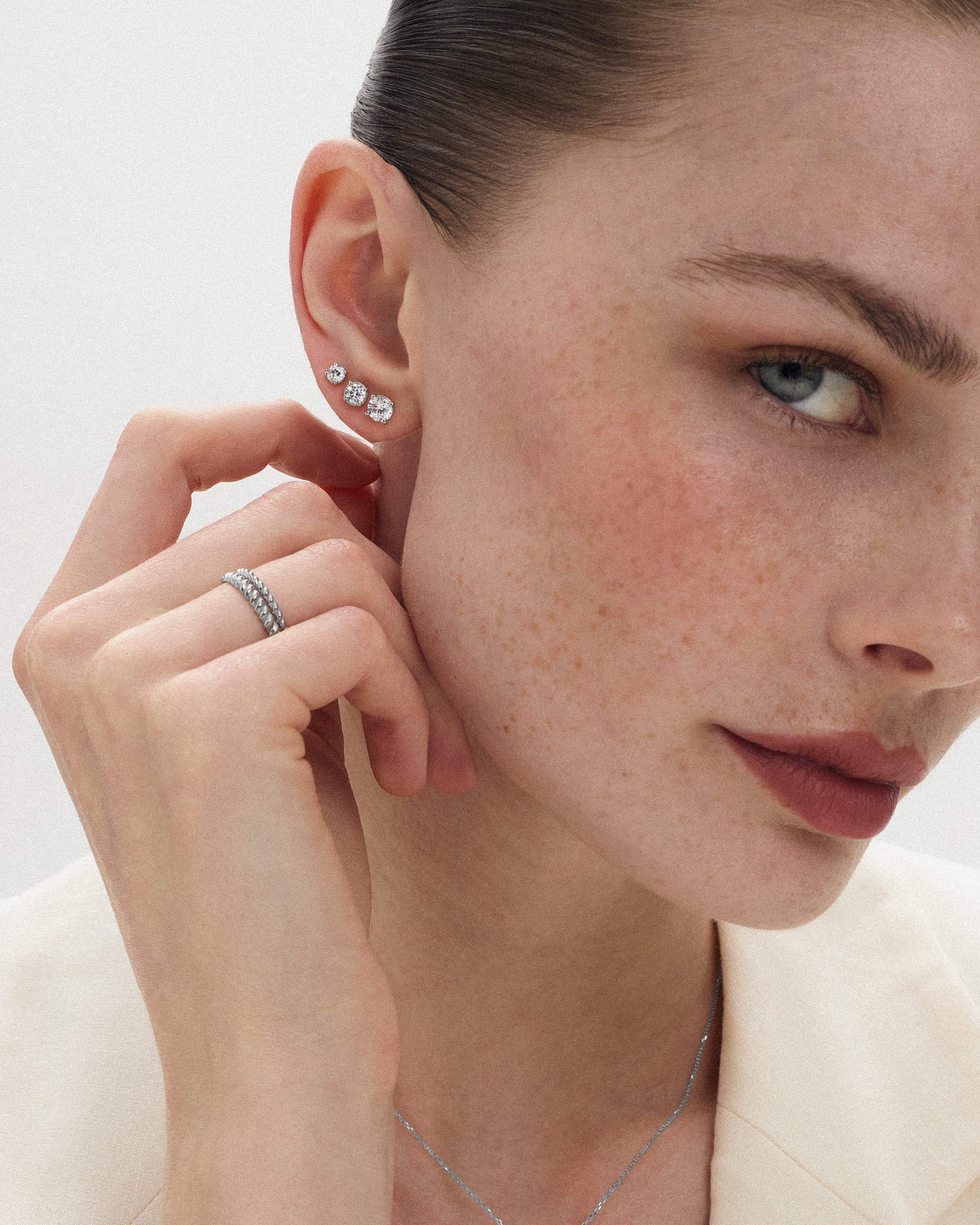 Angled view of a woman wearing 14k white gold bands alongside three eclat grand diamond studs of slightly different sizes with her hand touching her neck against a soft light background