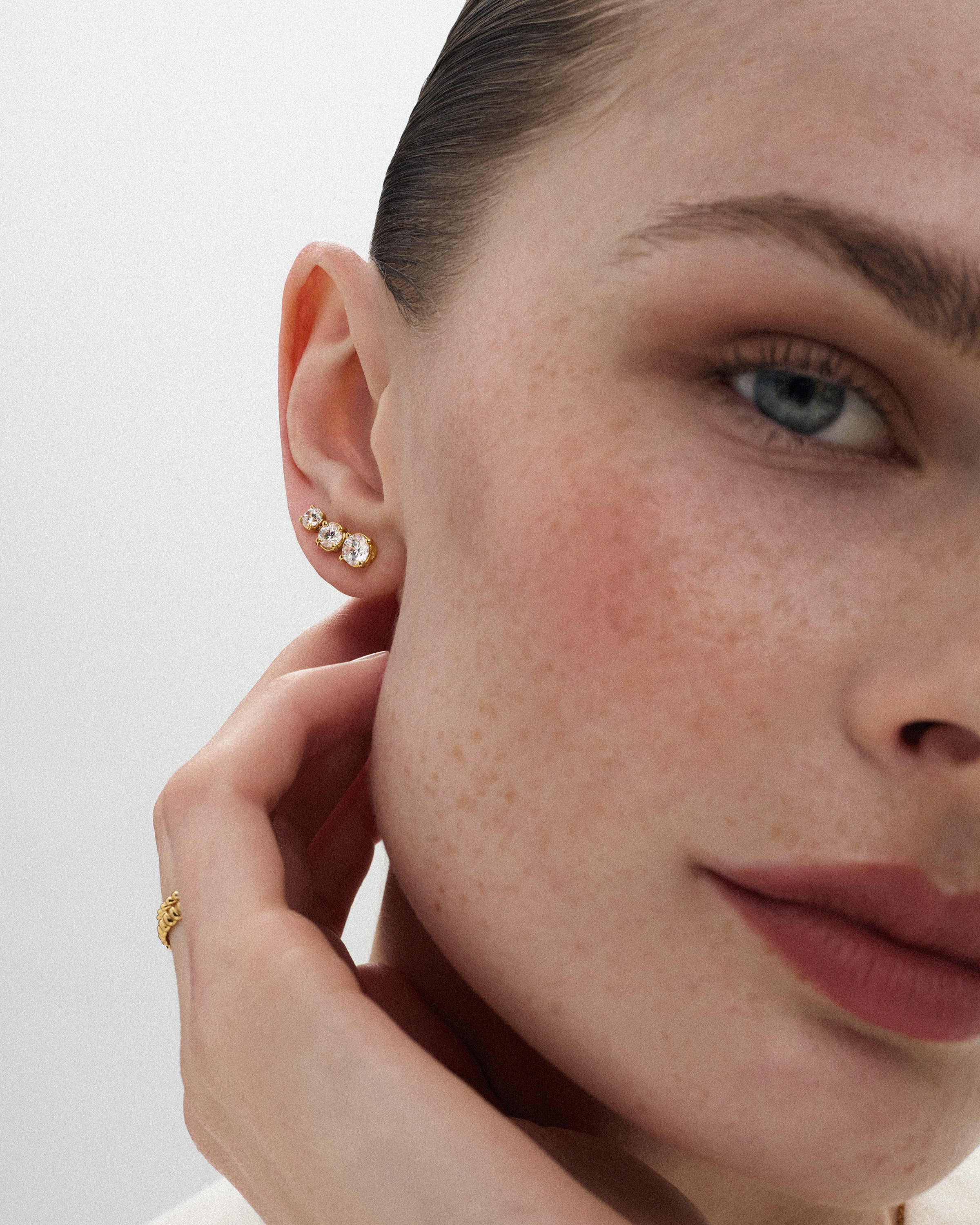 Angled view of a woman wearing three eclat grand stud earrings of different diamond sizes in 14k yellow gold against a light off-white background.