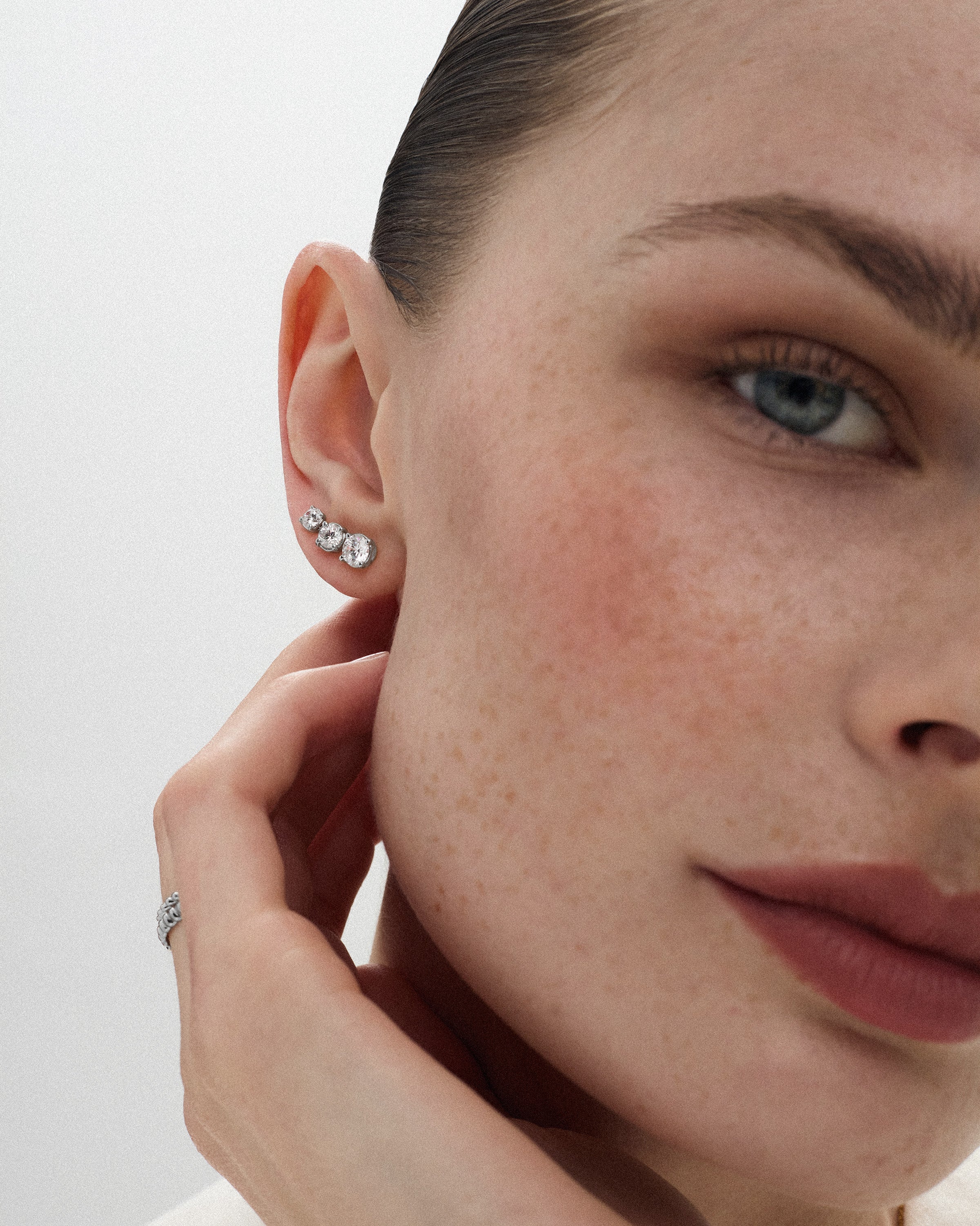 Angled view of a woman wearing three eclat grand stud earrings of different diamond sizes in 14k white gold against a light off-white background.