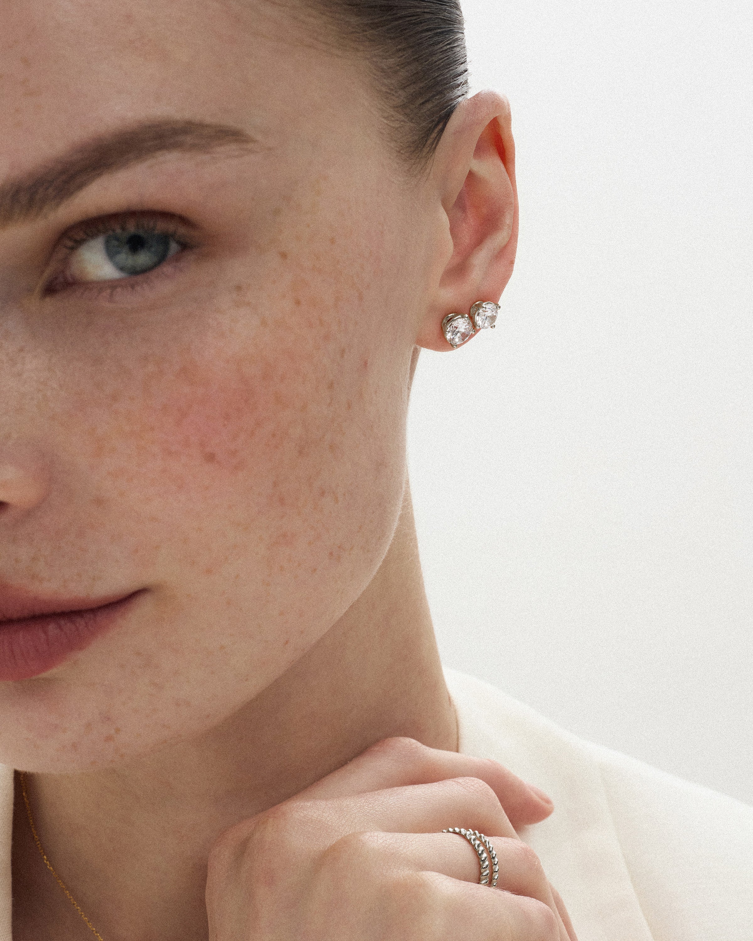 Angled view of a woman wearing two eclat grand stud earrings of different diamond sizes in 14k white gold against a light off-white background.