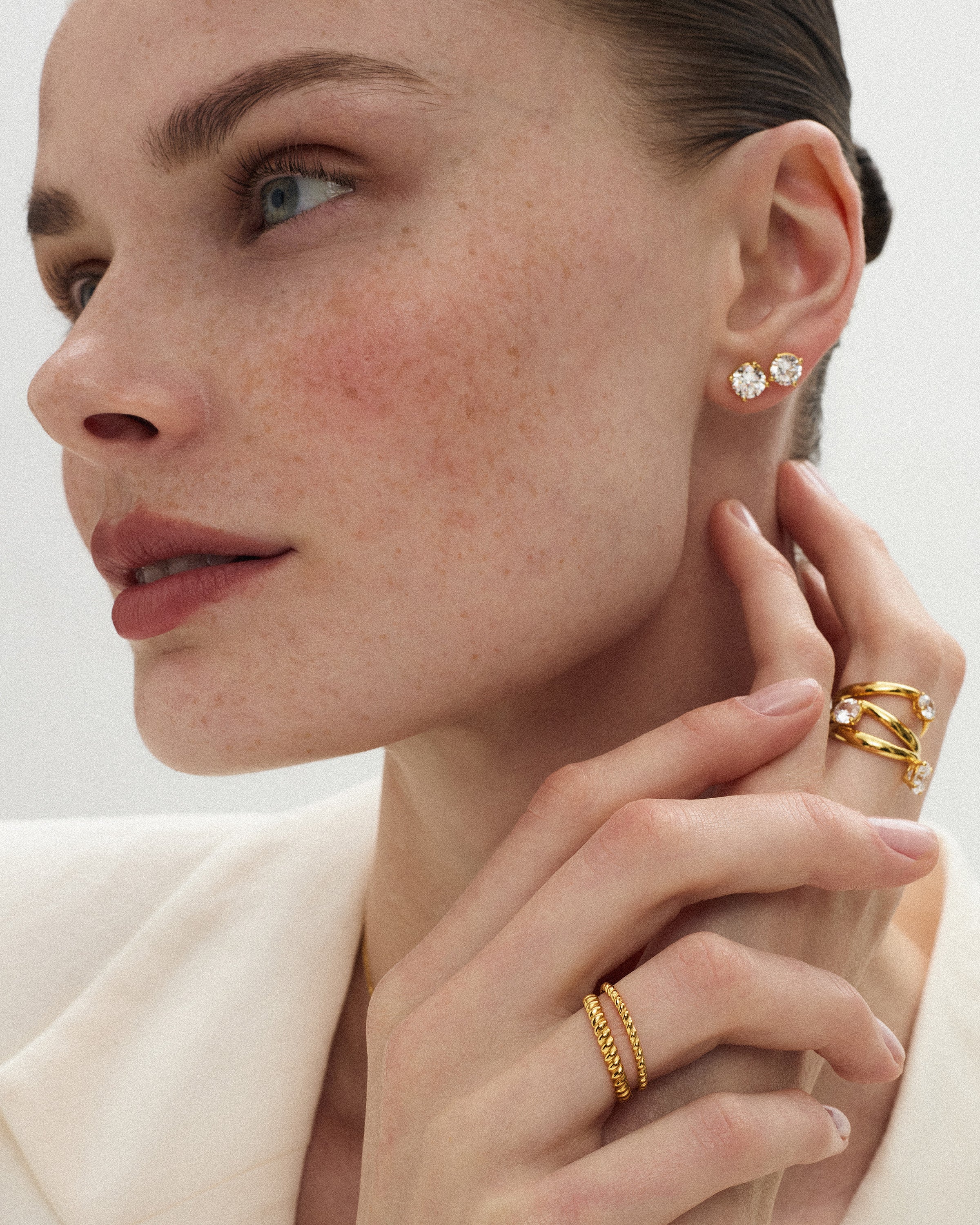 Close-up of a woman wearing 14k yellow gold solitaire rings and thin twisted bands alongside two eclat grand diamond studs of slightly different sizes with her hand near her neck against a soft light background.