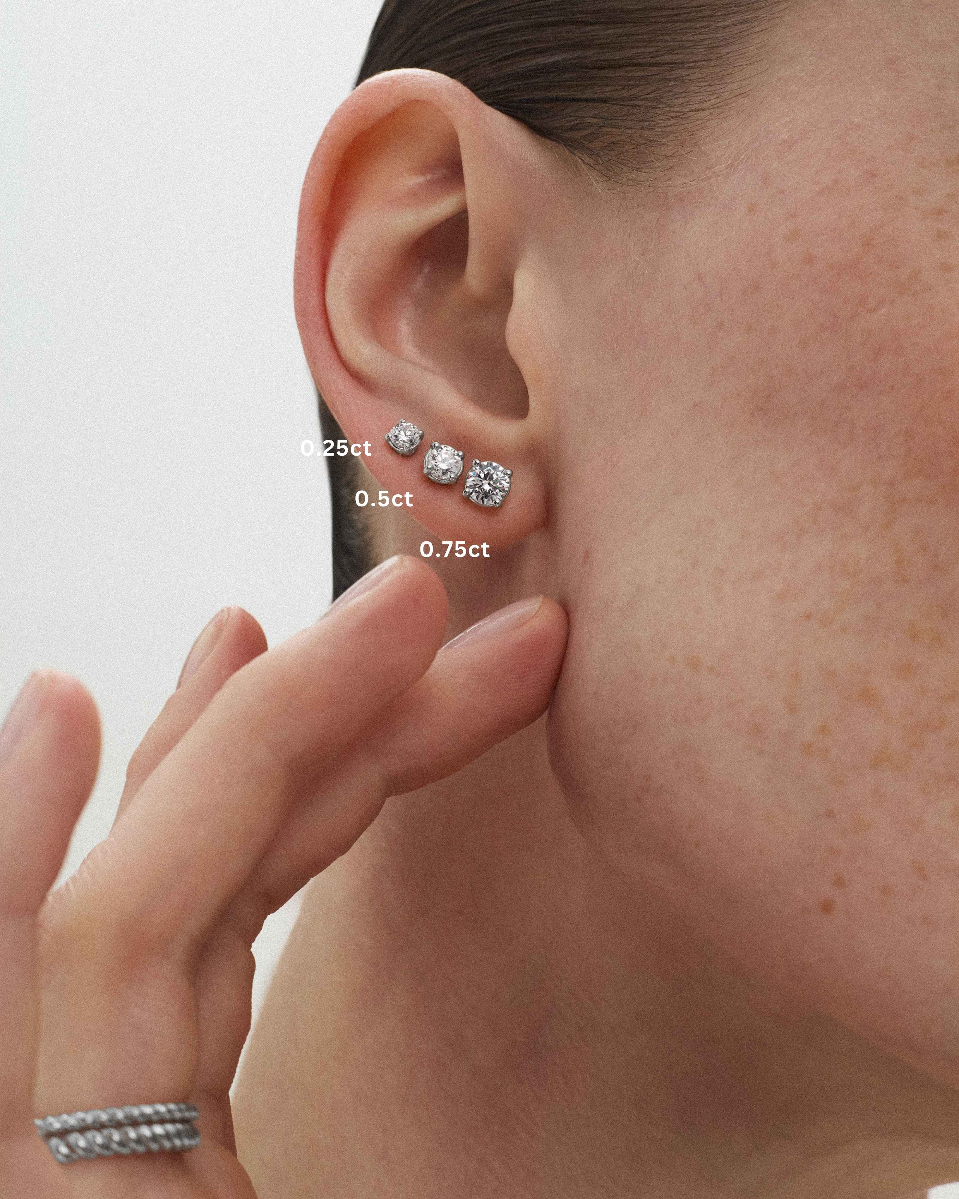A close-up of a woman's ear wearing three eclat grand stud earrings in 14k white gold with the diamond sizes 0.25ct, 0.5ct, and 0.75ct being indicated against a light off-white background