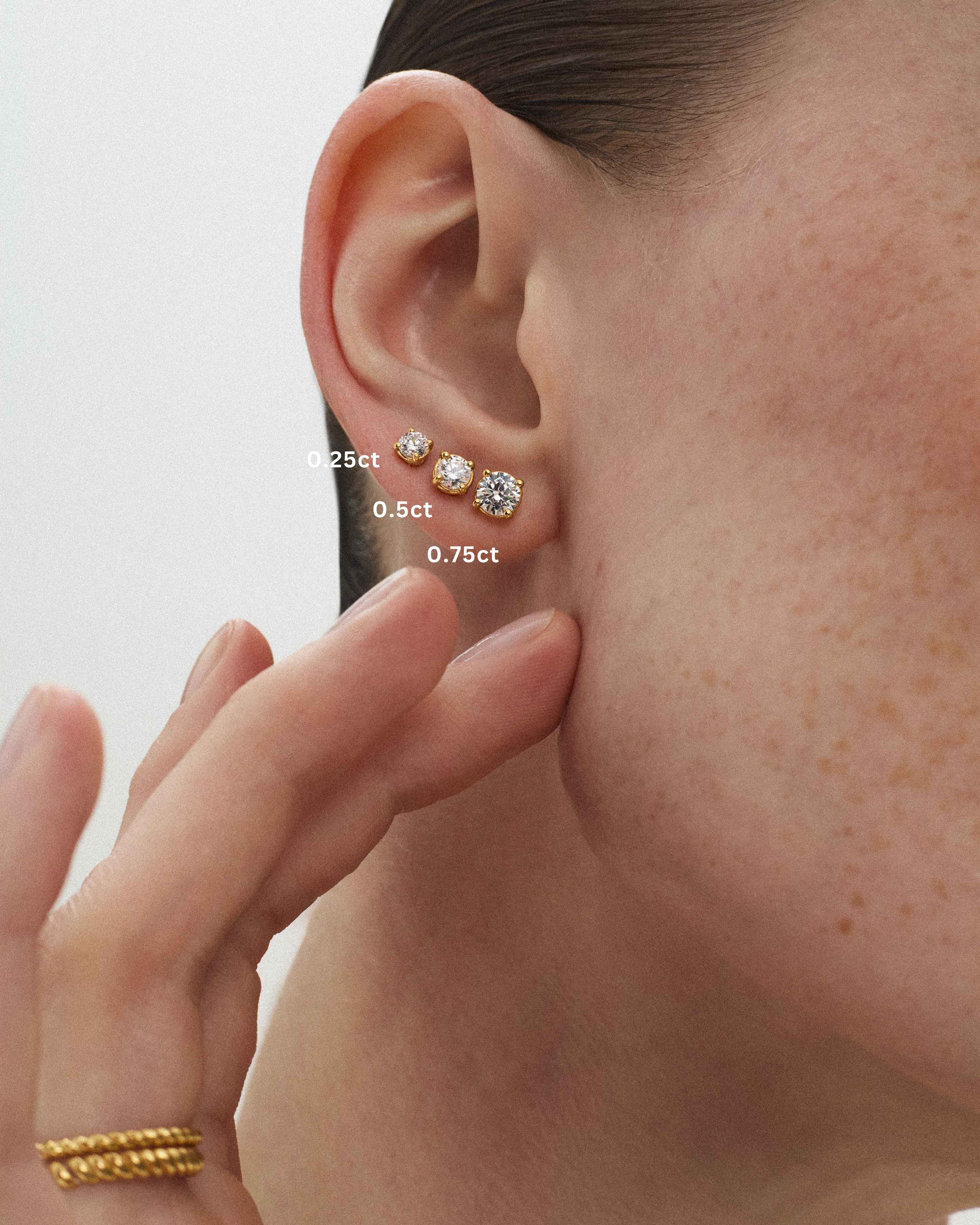 A close-up of a woman's ear wearing three eclat grand stud earrings in 14k yellow gold with the diamond sizes 0.25ct, 0.5ct, and 0.75ct being indicated against a light off-white background