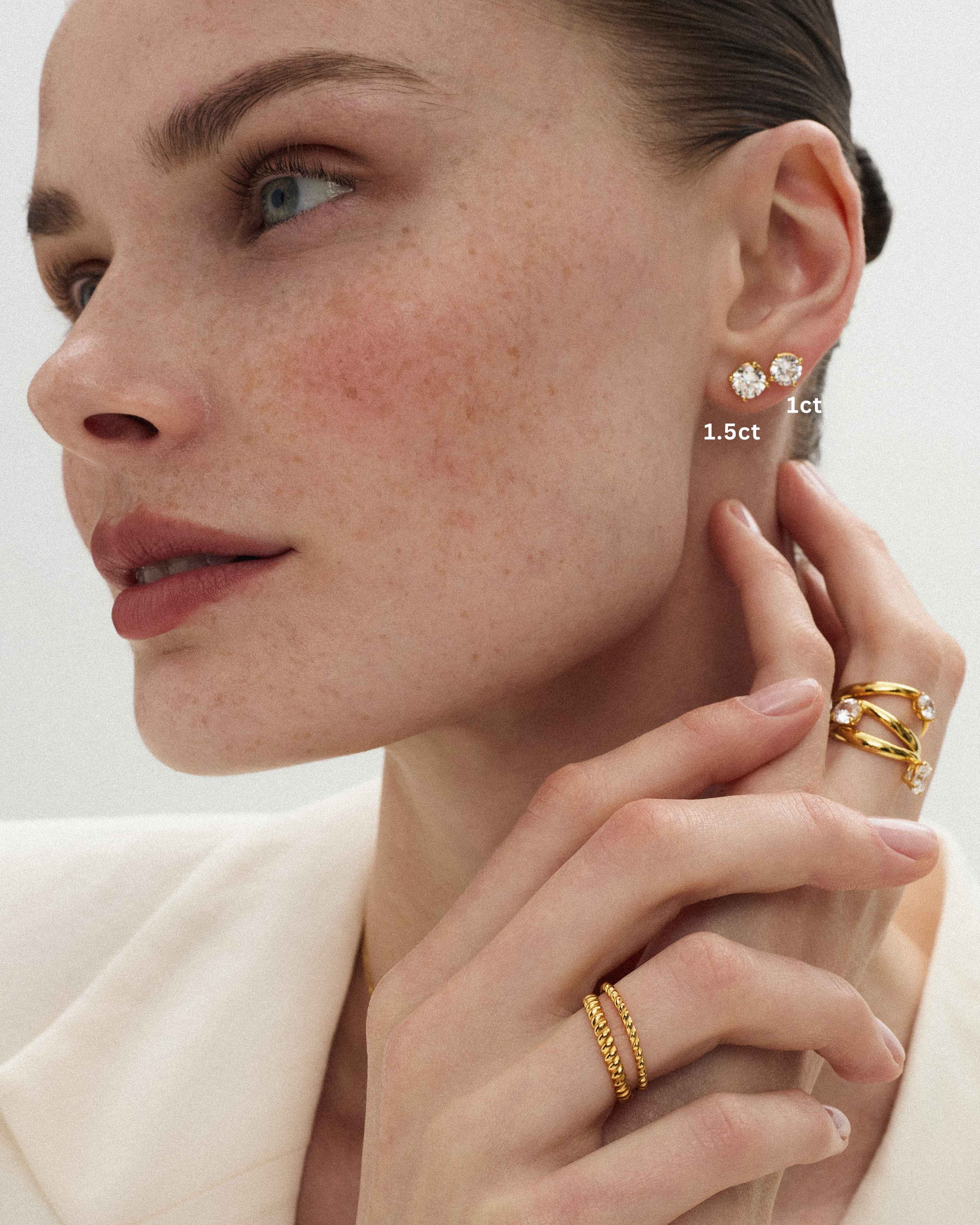 Close-up of a woman wearing 14k yellow gold solitaire rings and thin twisted bands alongside two eclat grand diamond studs of 1ct and 1.5ct diamond sizes stacked on her ear with her hand near her neck against a soft light background.