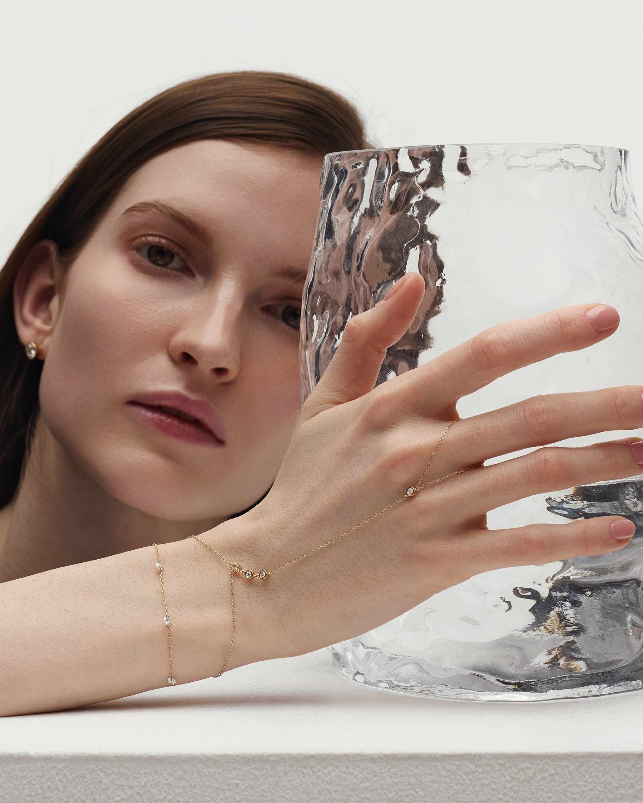 woman standing behind a glass object holding it with her hand styled with a solid gold chain bracelet adorned with small bezel diamonds and a delicate diamond station bracelet