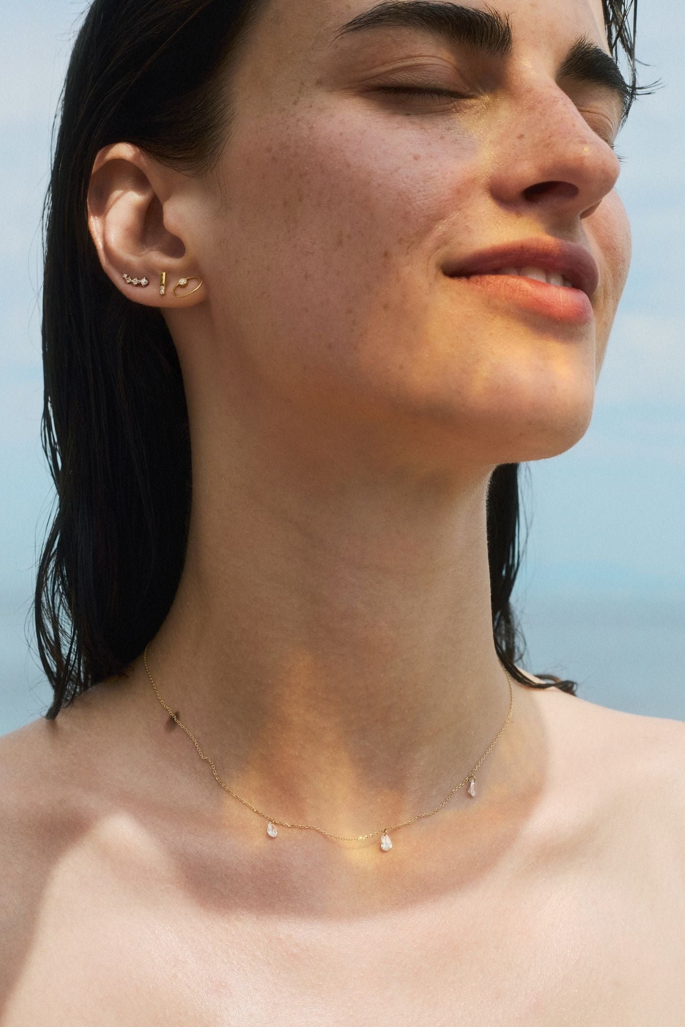 A close-up shot of a woman with closed eyes under soft natural light, wearing a delicate gold chain necklace with three dangling pear-shaped diamonds and dainty gold hoop earrings against a blurred sea background.